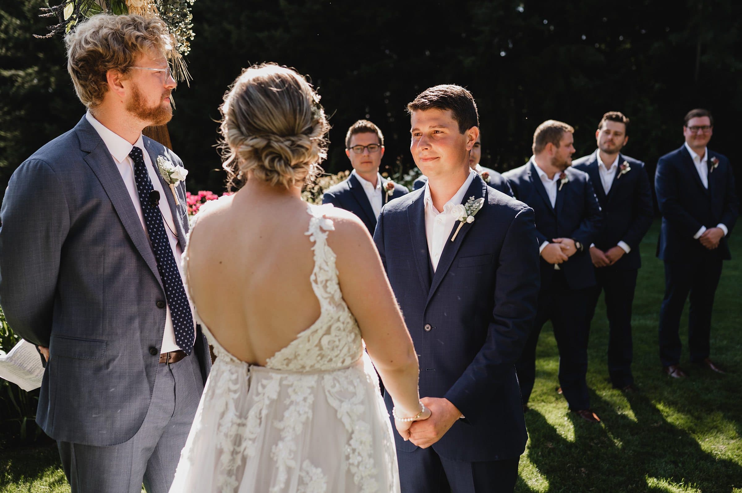 A couple holding hands during their outdoor wedding ceremony at Gorge Crest Vineyards, with attendants standing nearby.