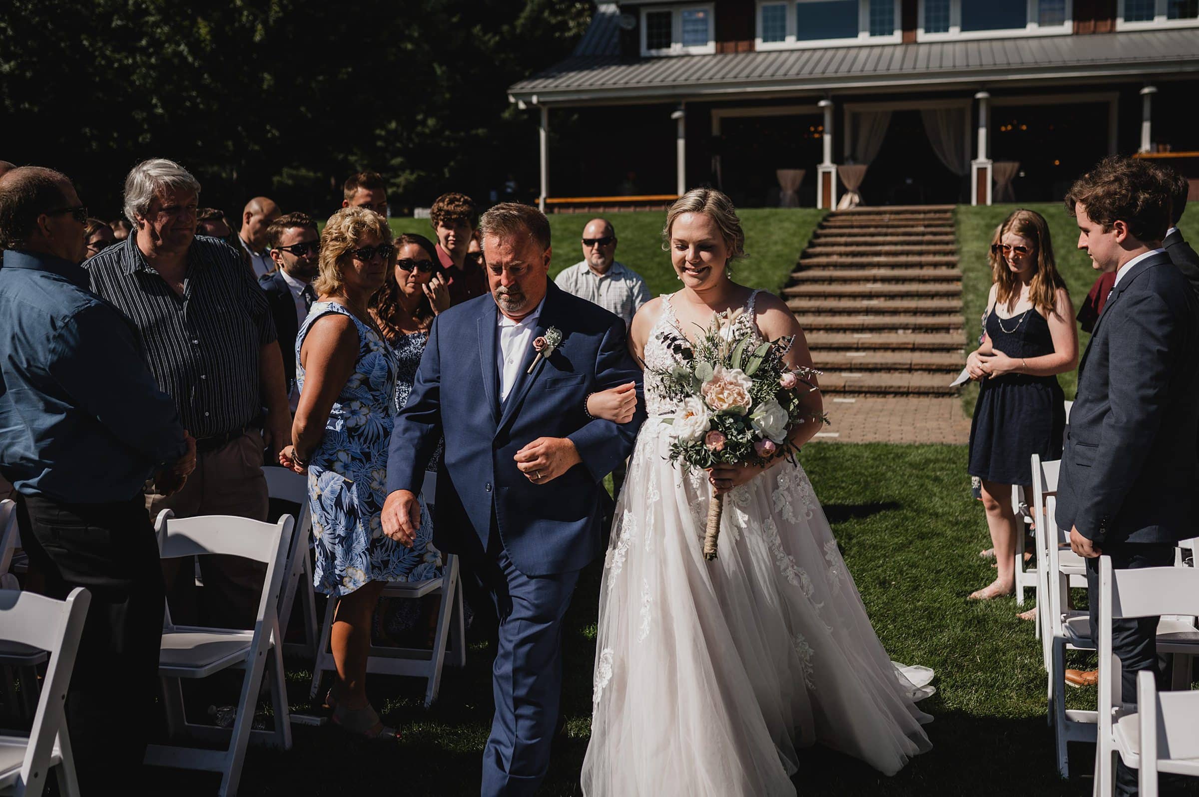 The bride in a white gown walks down the aisle with a man in a suit, surrounded by guests outdoors at Gorge Crest Vineyards, where they capture every precious moment with stunning wedding photography in front of the elegant building.