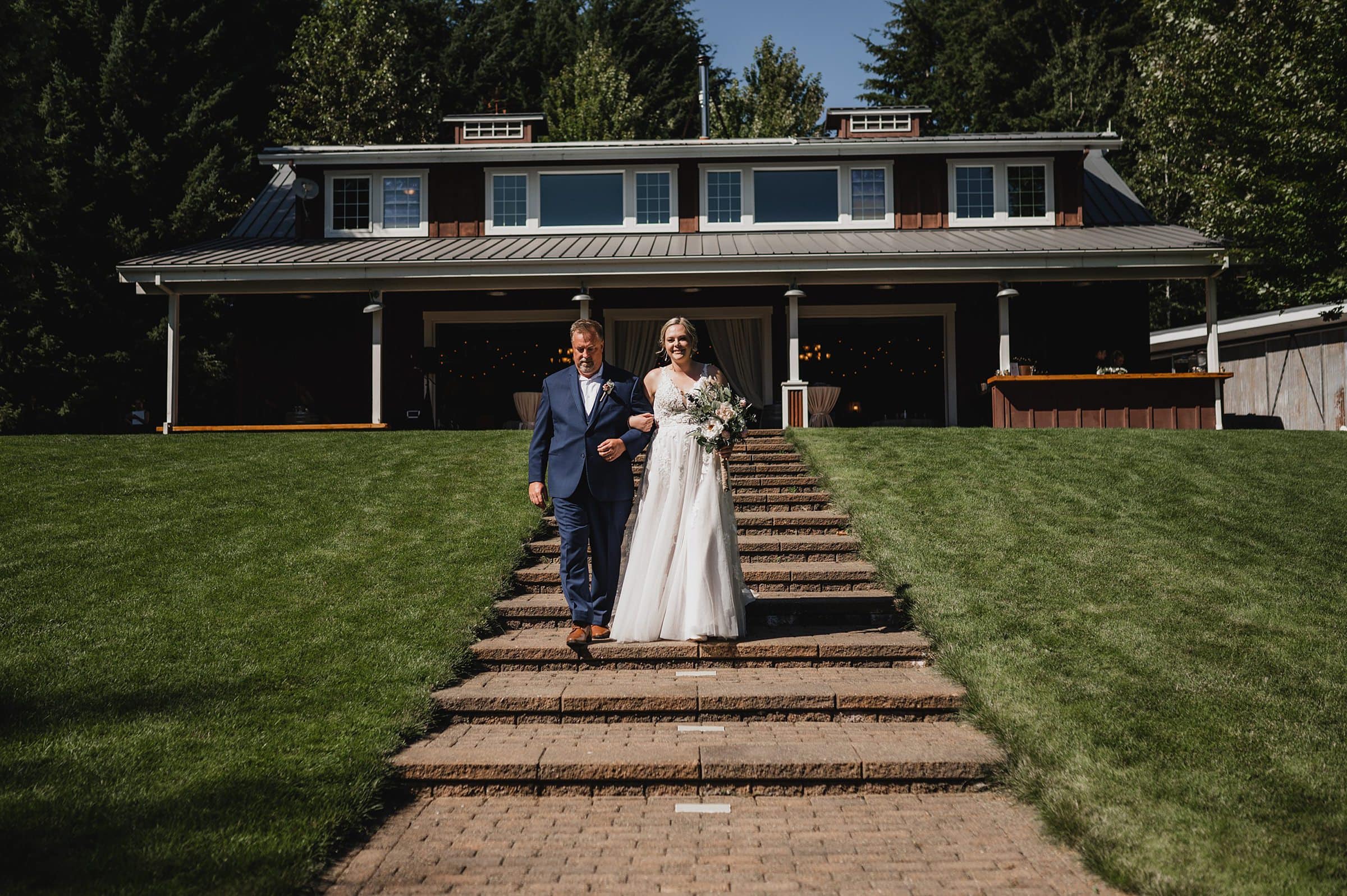 A bride and groom descend outdoor steps in front of a large house, surrounded by lush greenery. This beautiful moment is captured at the stunning Gorge Crest Vineyards, offering memorable wedding photography for couples.