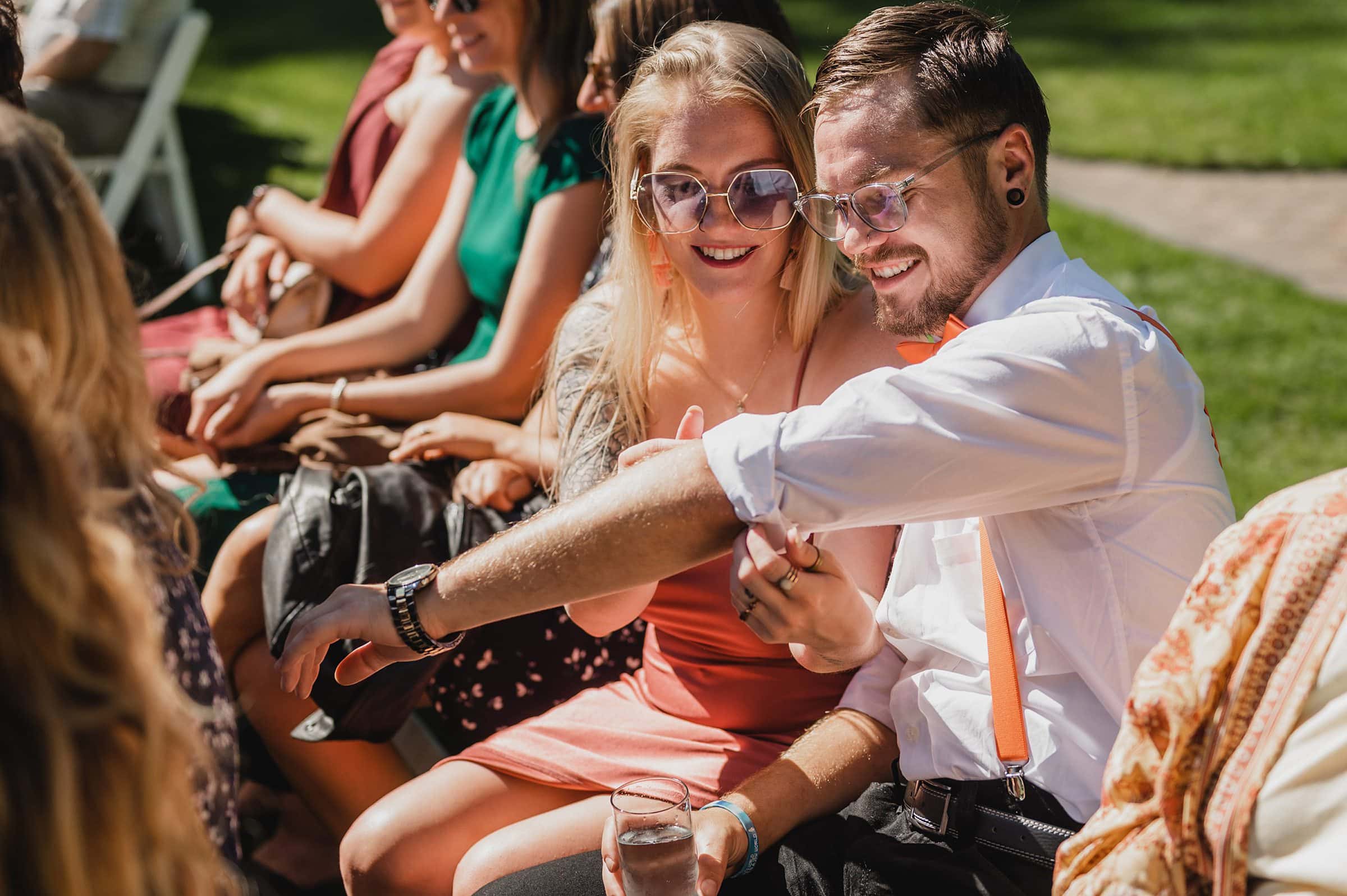 A smiling couple seated outdoors, with the woman wearing sunglasses and the man in a shirt with suspenders, capture a joyful moment at Gorge Crest Vineyards.
