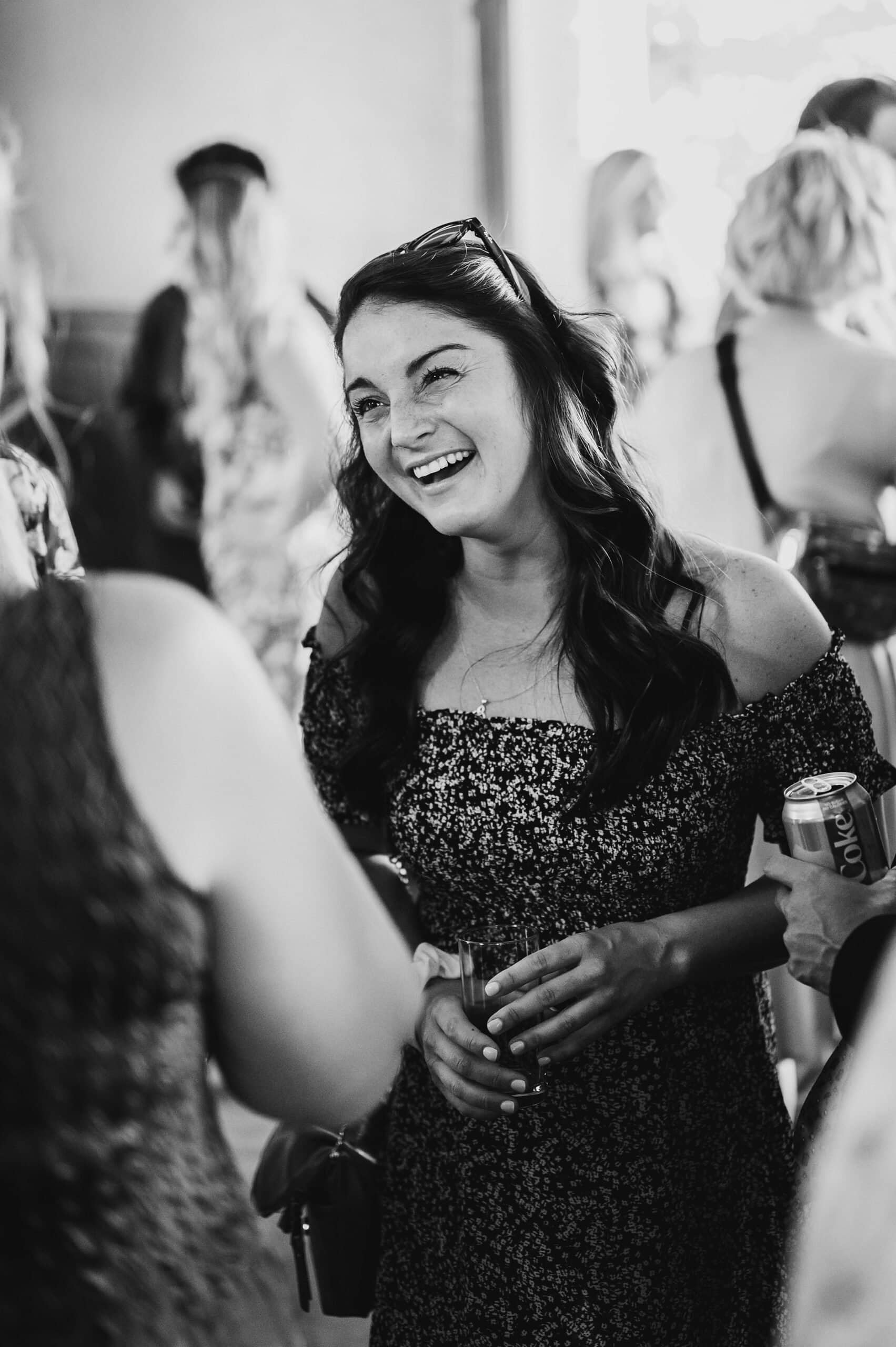 A woman smiles and chats at a social gathering, elegantly holding a can and cup. Set against the timeless backdrop of Gorge Crest Vineyards, this black-and-white photo captures the candid joy often found in wedding celebrations.