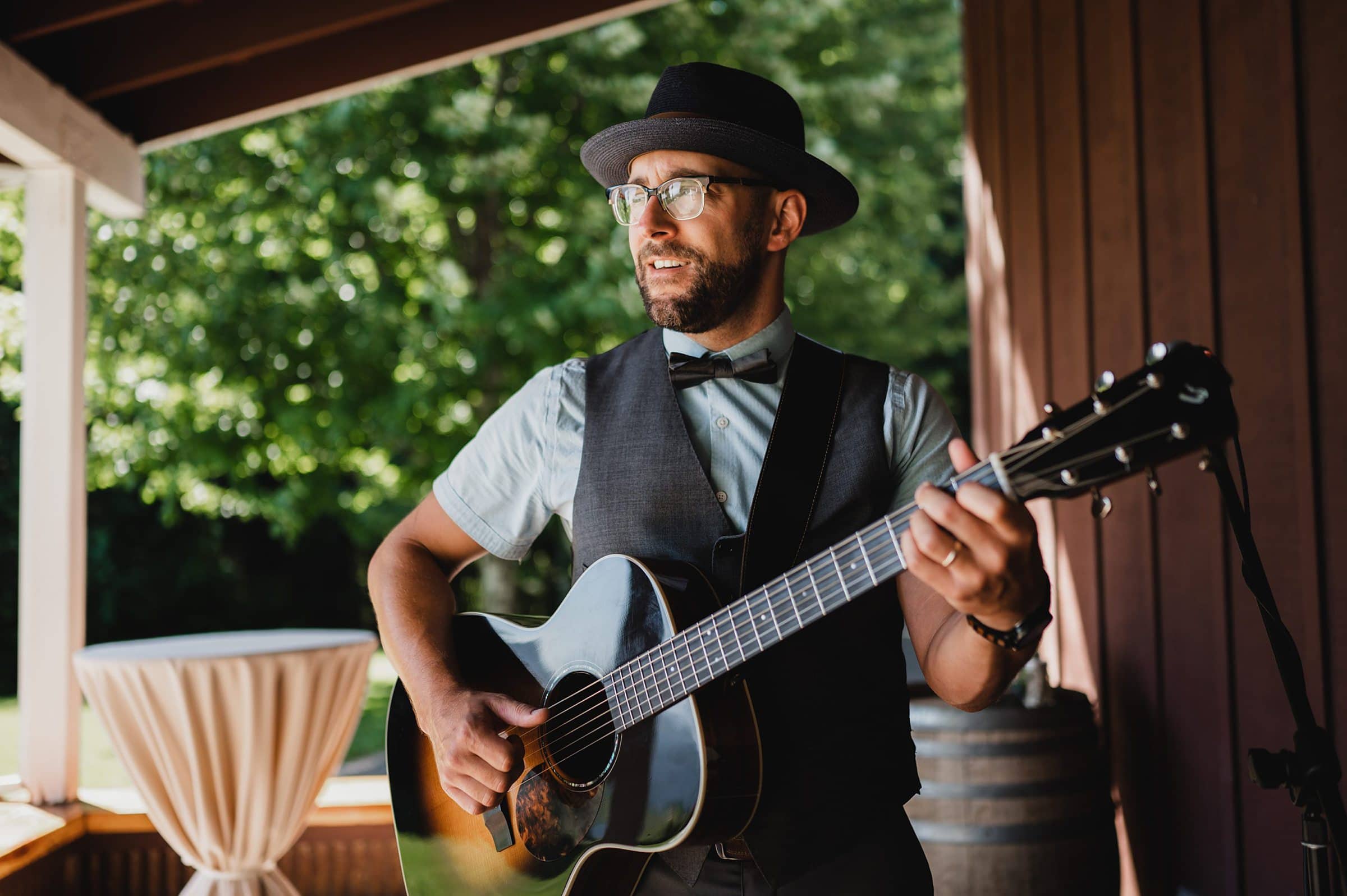 A man in a hat and vest plays an acoustic guitar outdoors by a wooden wall, surrounded by trees. Perfect for capturing breathtaking Gorge Crest Vineyards wedding photos.