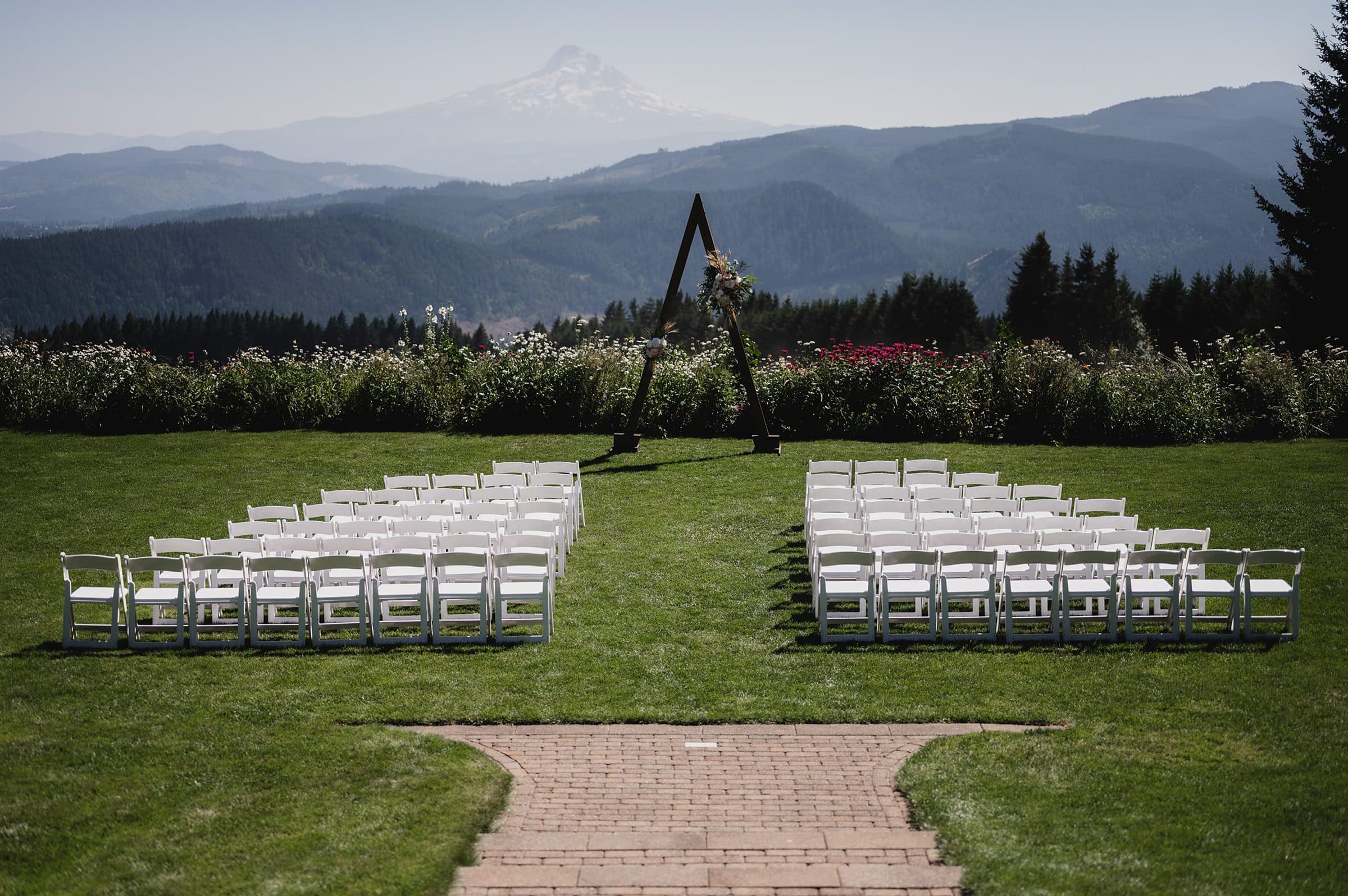 White chairs line the grassy field beneath a striking triangular arch, set against the breathtaking mountain backdrop of Gorge Crest Vineyards. Perfect for capturing stunning wedding photos, this venue offers a majestic blend of natural beauty and elegance.