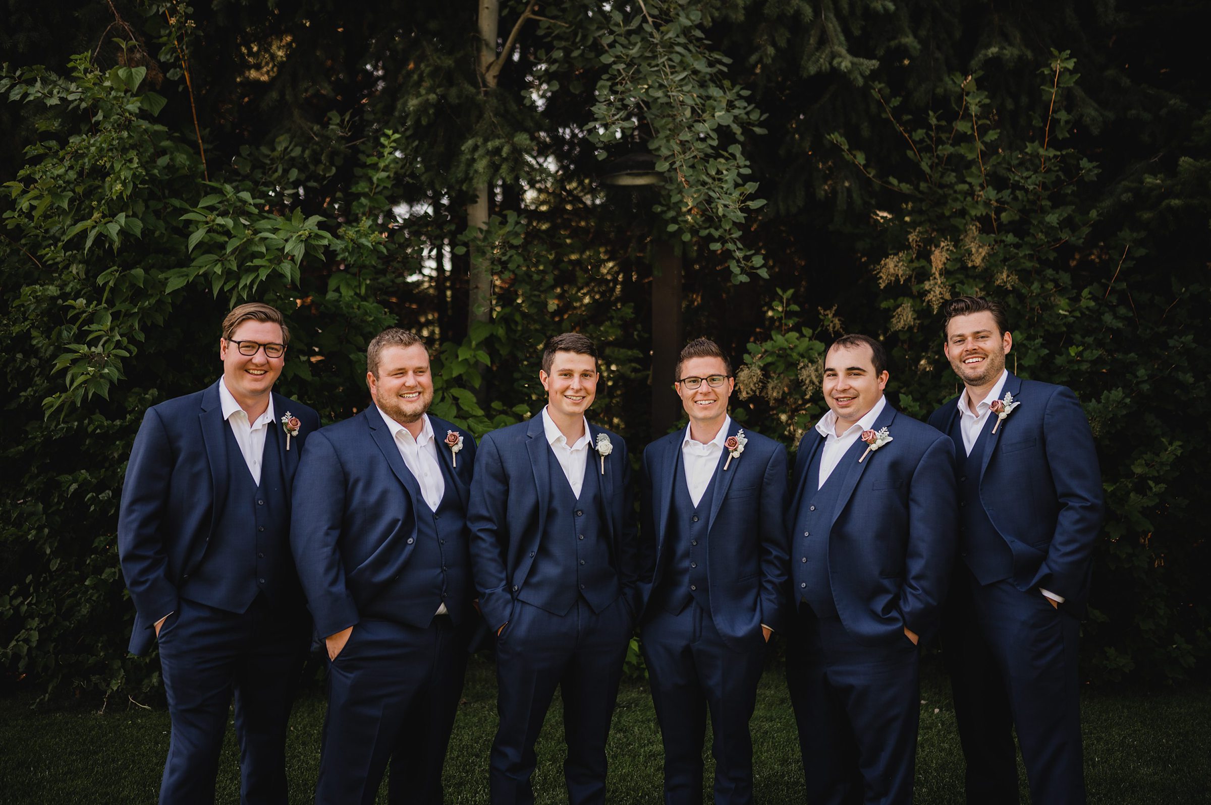 Six men in blue suits and boutonnières stand outdoors, smiling against a backdrop of lush greenery at Gorge Crest Vineyards.