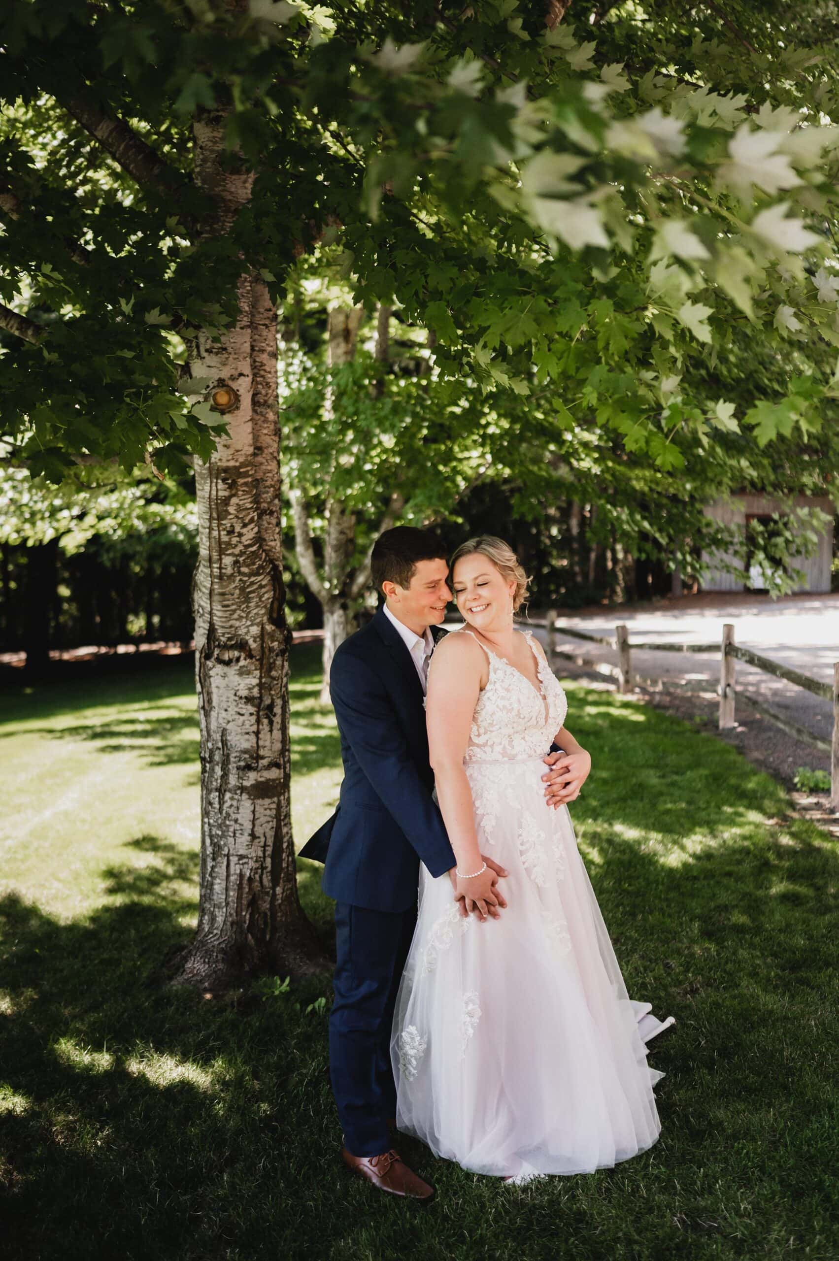 The bride and groom smile under a tree at Gorge Crest Vineyards, with sunlight filtering through the leaves onto the grassy area.