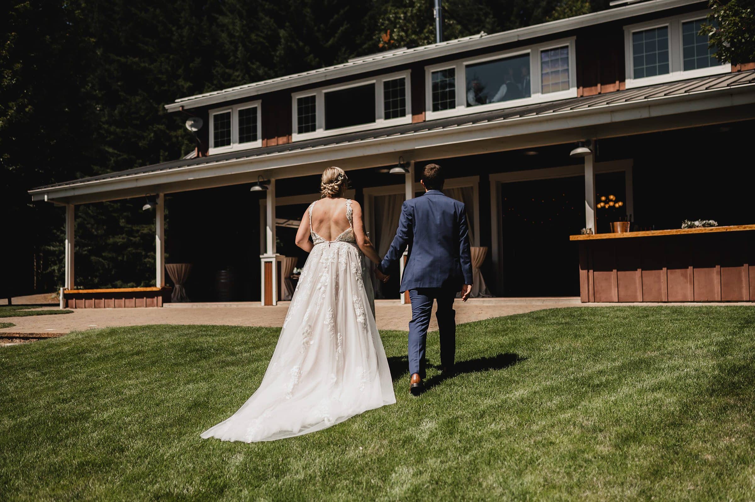 A bride and groom walk hand in hand on the grass toward a rustic building at Gorge Crest Vineyards, with picturesque scenery in the background.