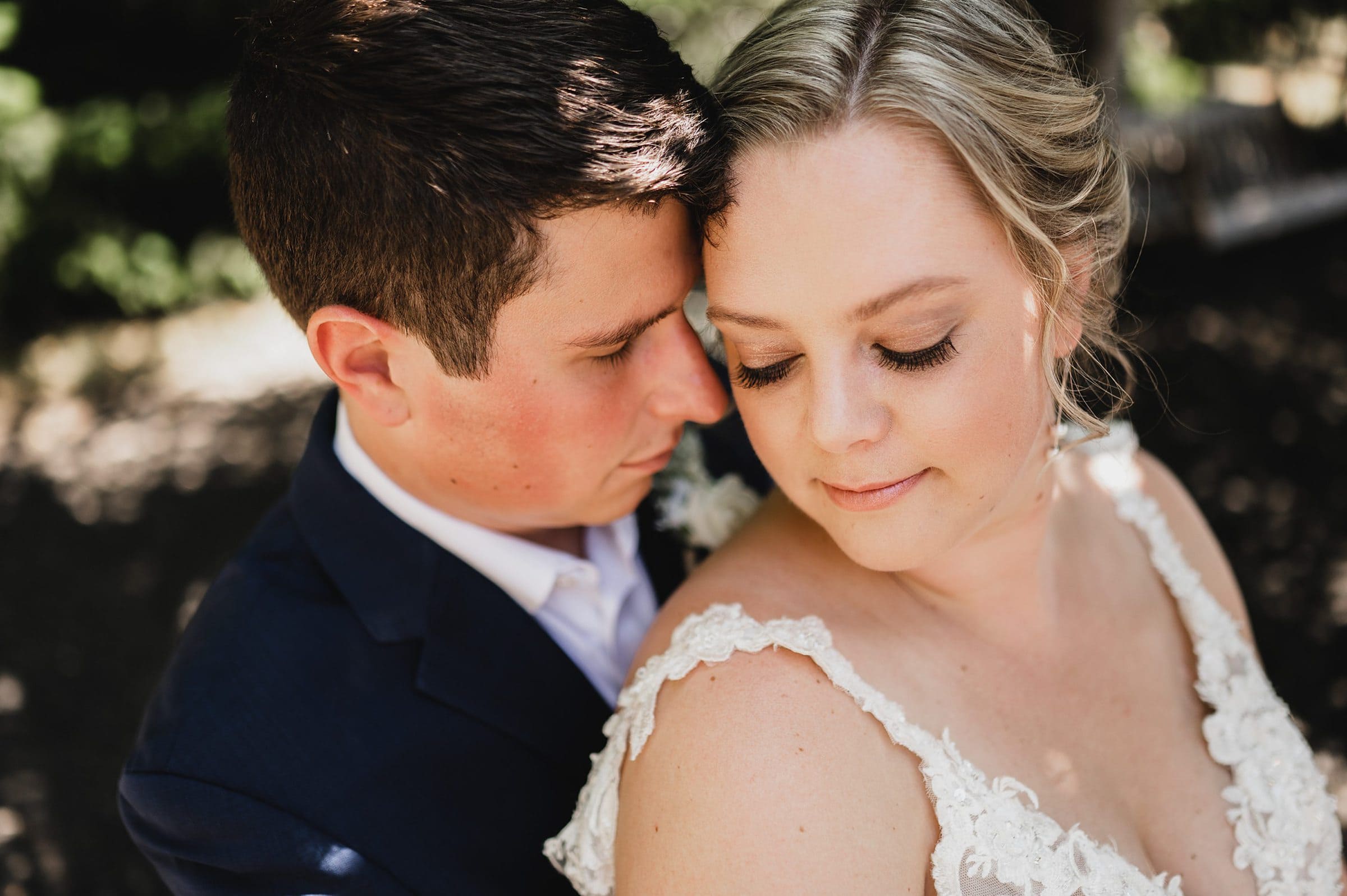 A couple embracing closely, the woman in a lace dress and the man in a dark suit, with eyes gently closed amidst a scenic vineyard backdrop.