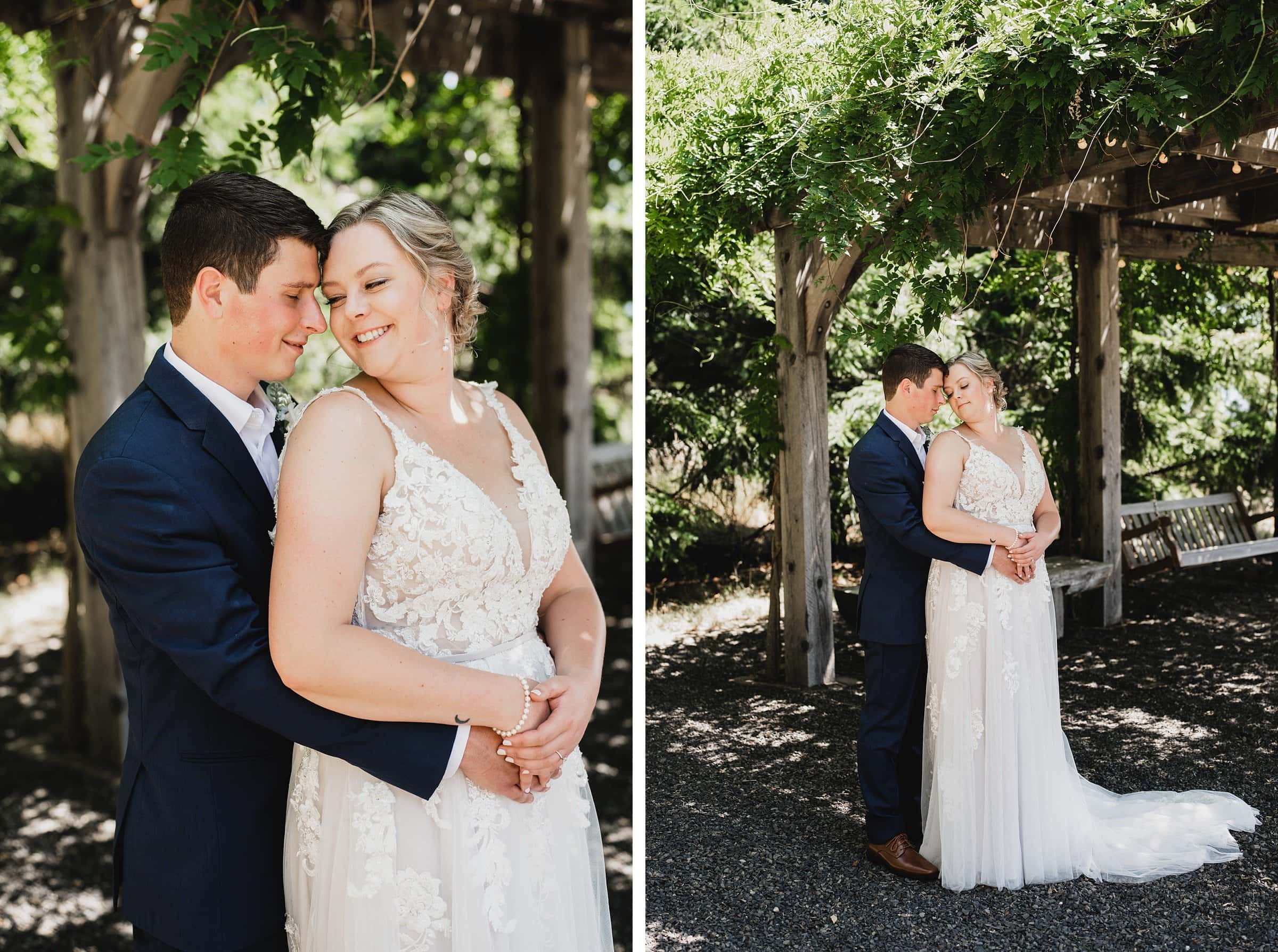 At Gorge Crest Vineyards, a couple in wedding attire embraces under a wooden pergola adorned with greenery, both smiling and standing closely together.