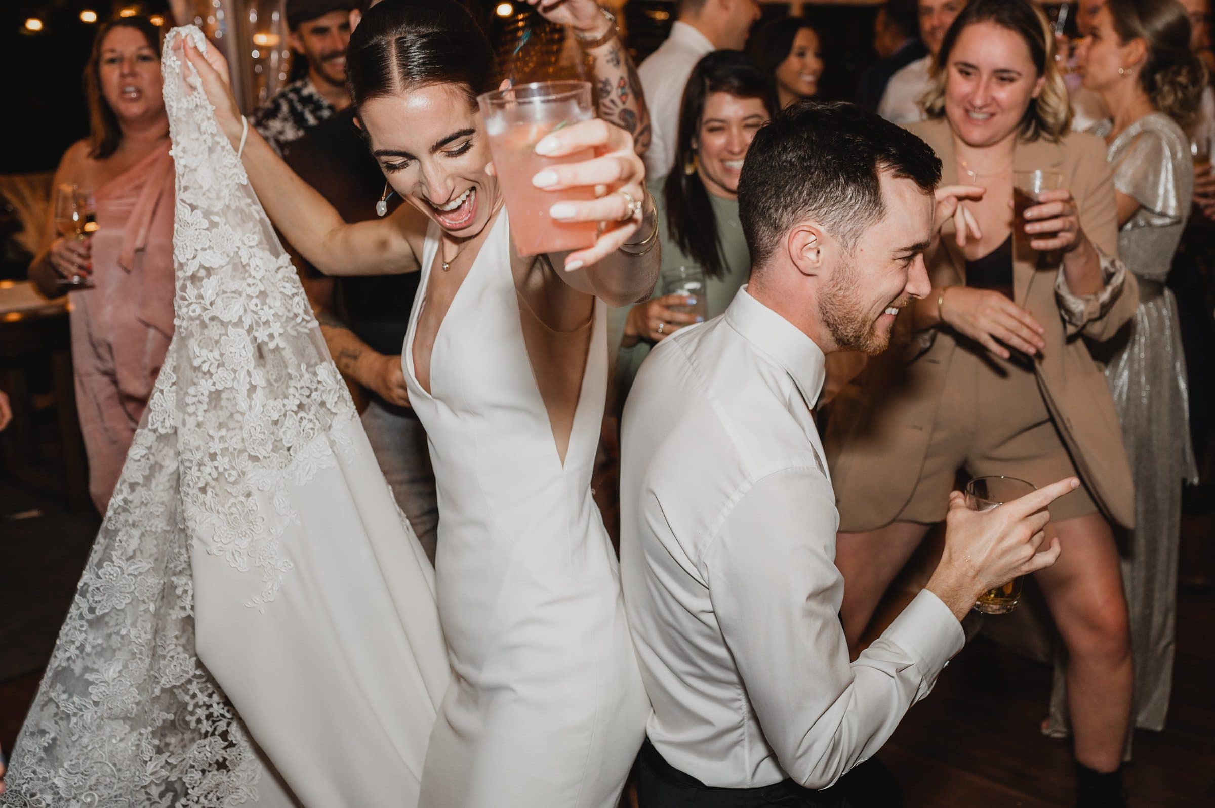 Bride and groom dancing joyfully with drinks, surrounded by a cheering crowd at a lively reception. Candid moments captured beautifully as part of their wedding photos at The Griffin House, preserving memories of an unforgettable celebration.