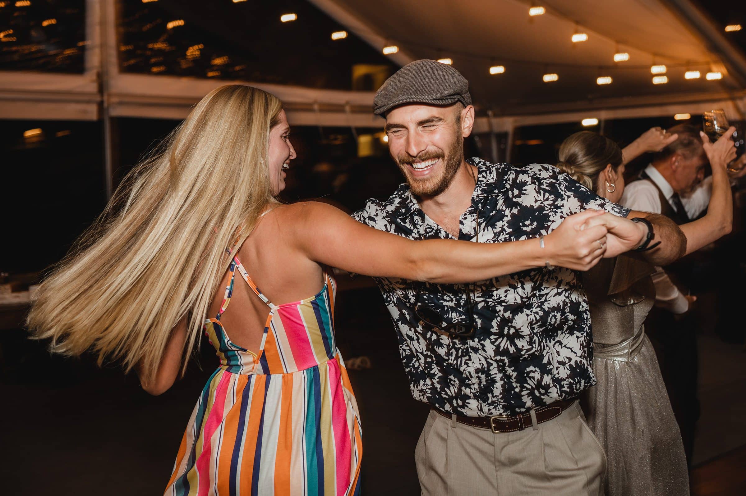 A man in a floral shirt and cap joyfully dances with a woman in a colorful dress at an outdoor evening wedding event, capturing the festive spirit in memorable photos.