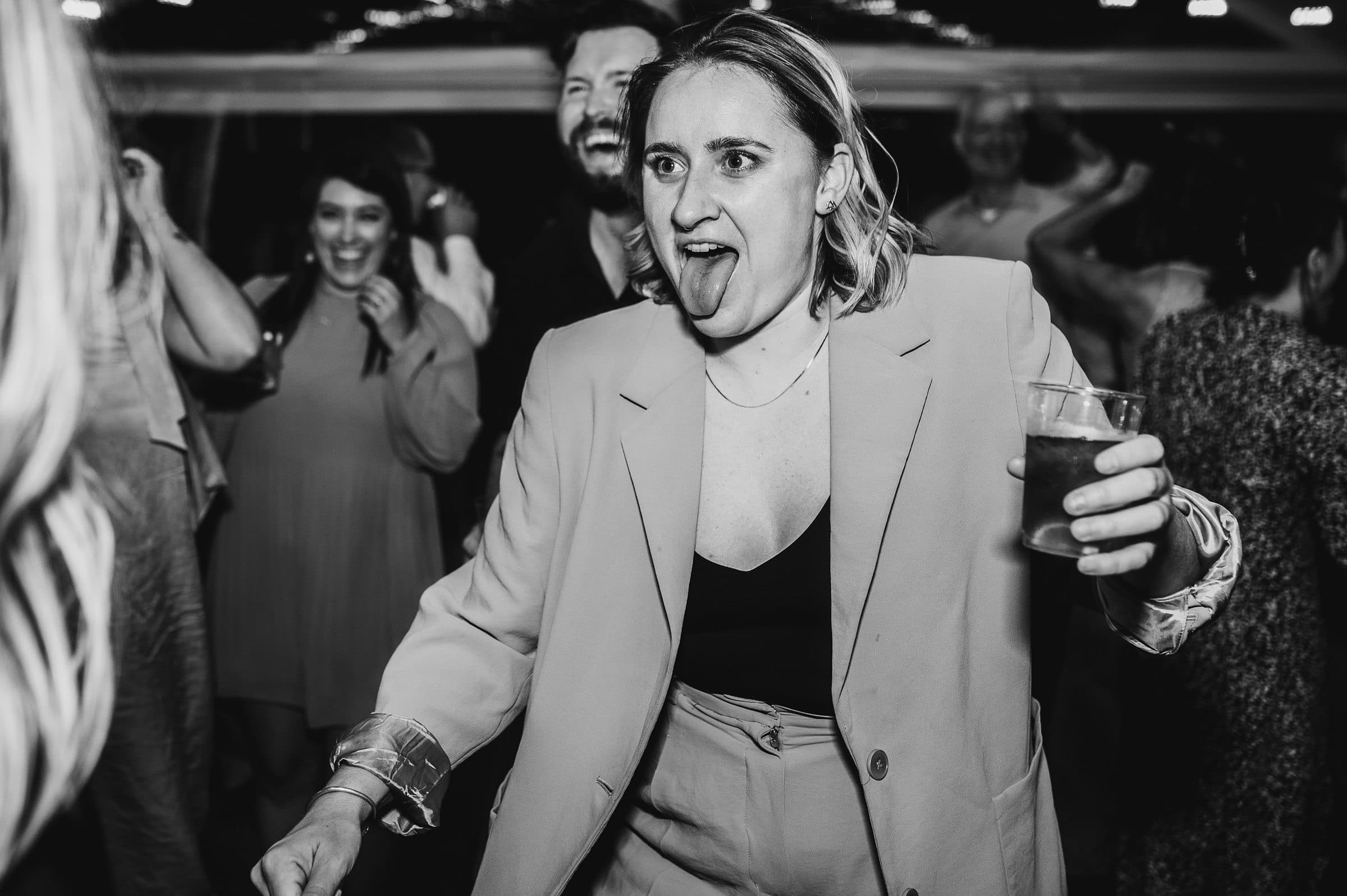 A person in a light-colored suit joyfully sticks out their tongue and holds a drink, enjoying wedding festivities at The Griffin House, with people dancing in the background.