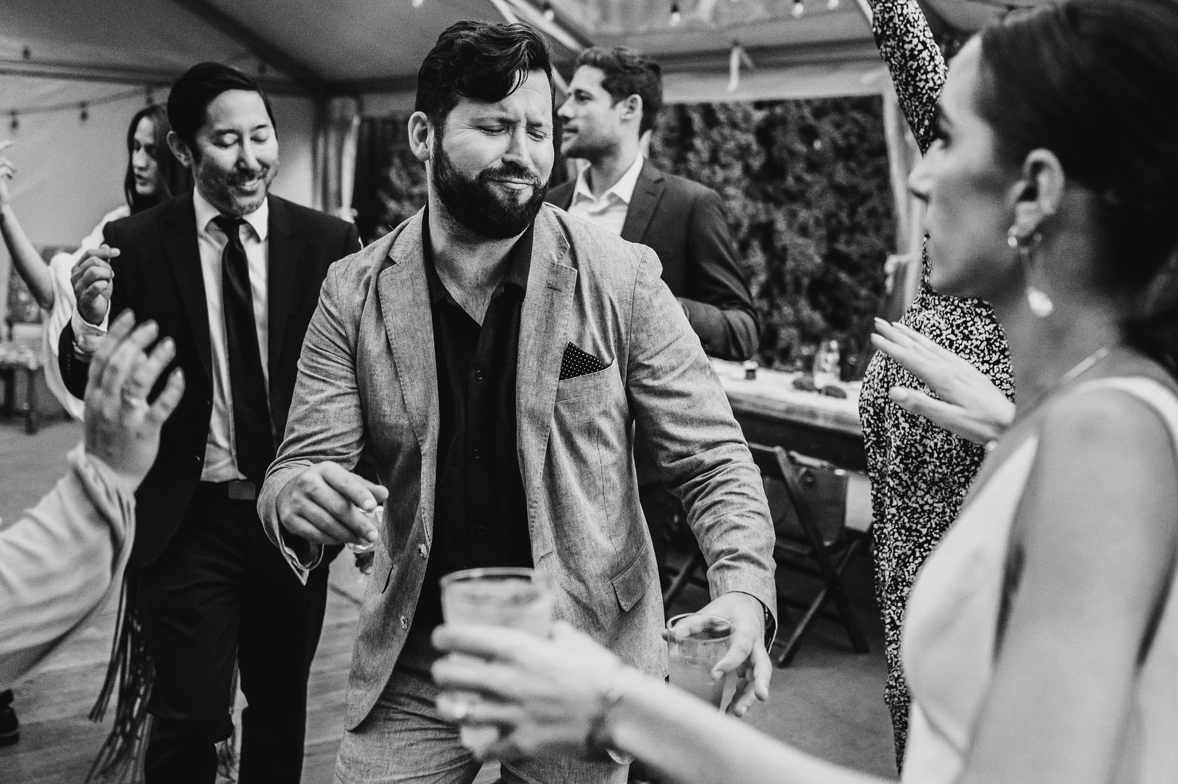 People in formal attire dancing energetically at a party, with one man in the center making a playful expression. The lively scene is akin to wedding photos at The Griffin House, capturing the joyful spirit and camaraderie of friends enjoying the celebration.