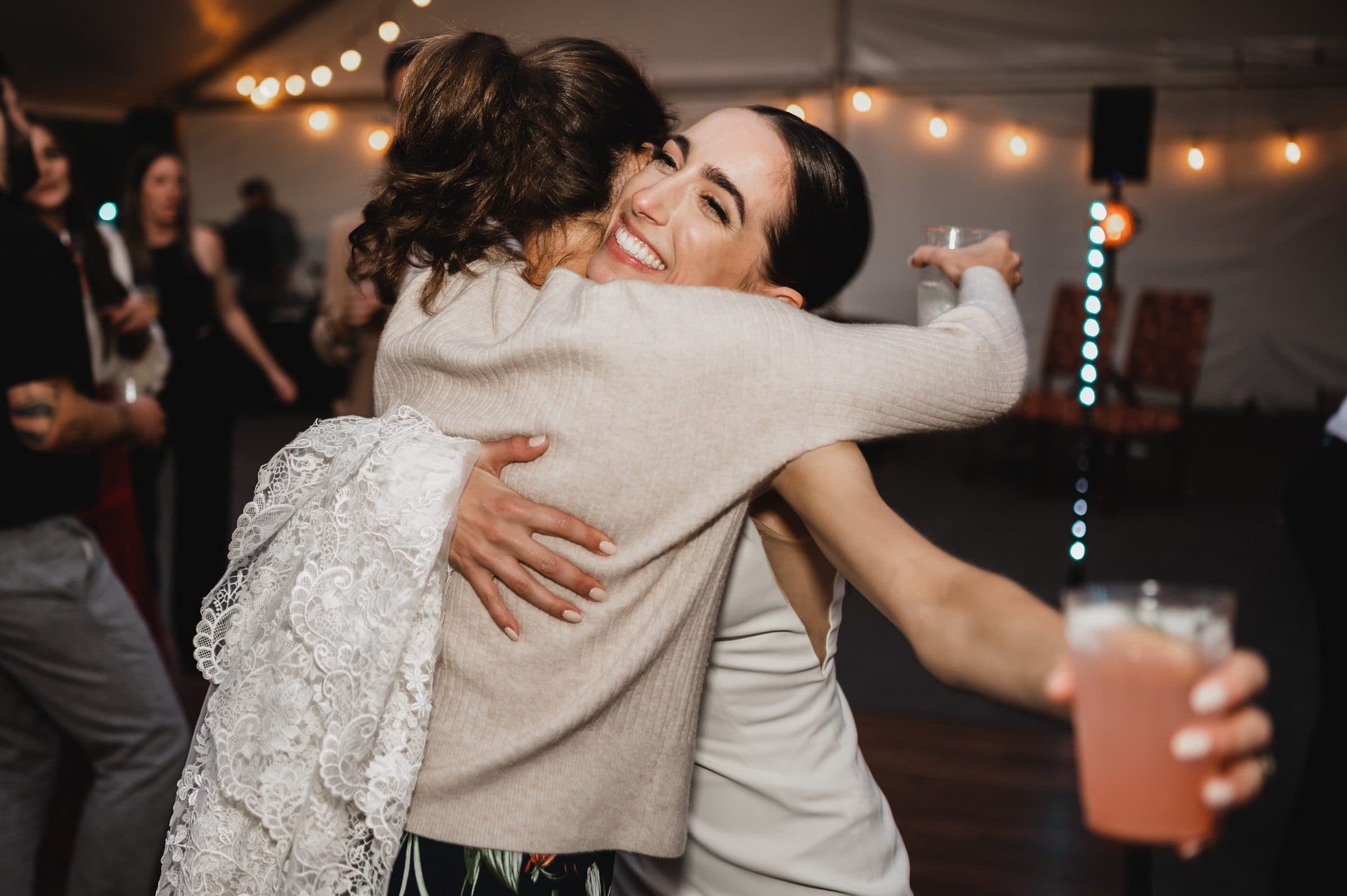 At a party illuminated by string lights, two people embrace joyfully, with one holding a drink. The scene resembles wedding photos at The Griffin House, capturing pure celebration and connection.