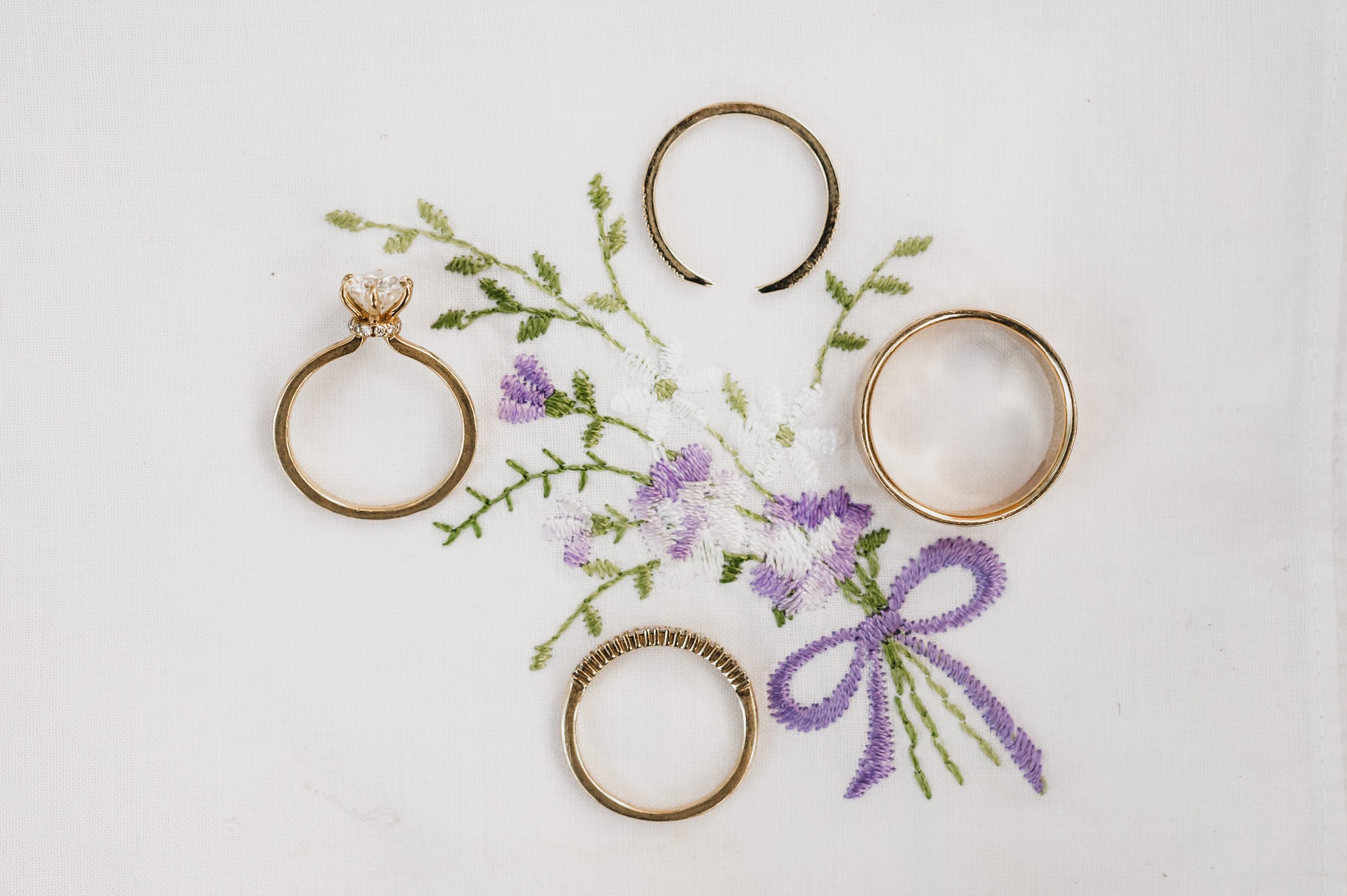 Four gold rings arranged in a circle on floral embroidery with purple flowers and a bow on white fabric create an elegant scene, perfect for wedding photos at The Griffin House.