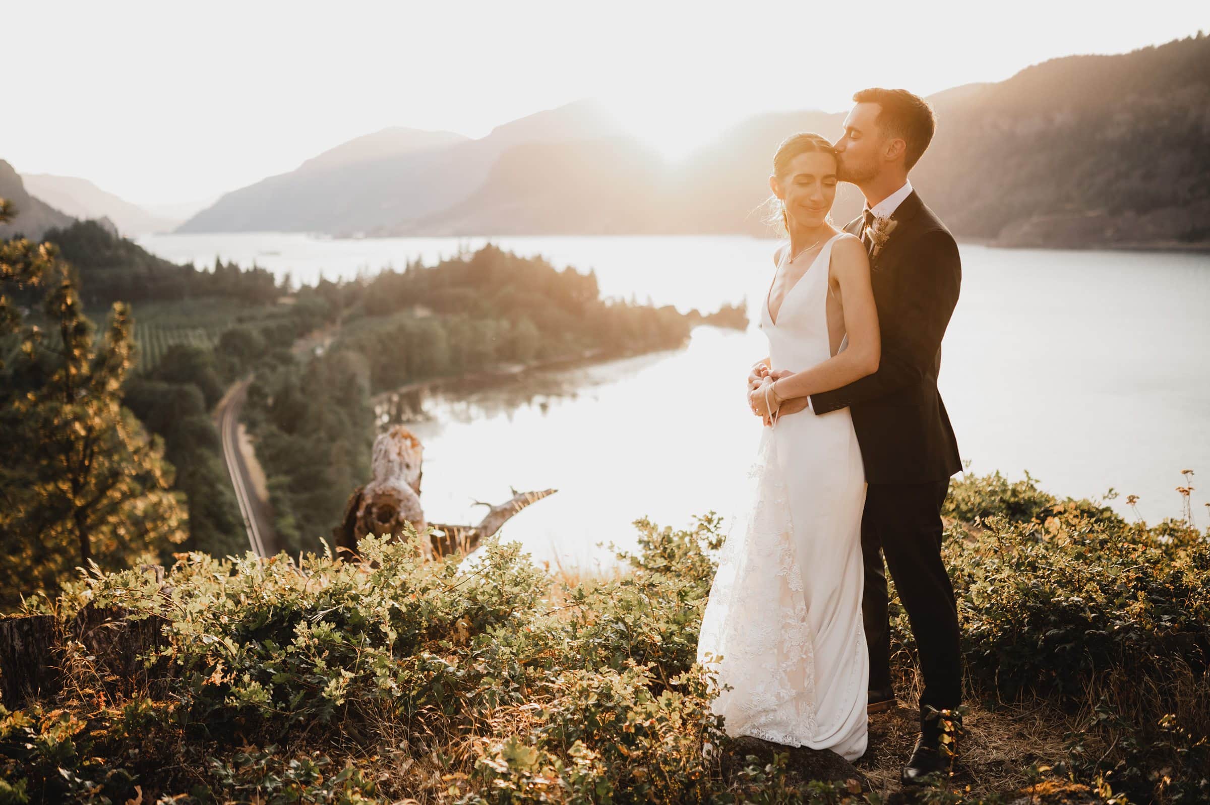 Bride and groom embrace on a scenic cliffside at sunset, overlooking the Columbia River Gorge and mountains, capturing perfect wedding photos at The Griffin House.