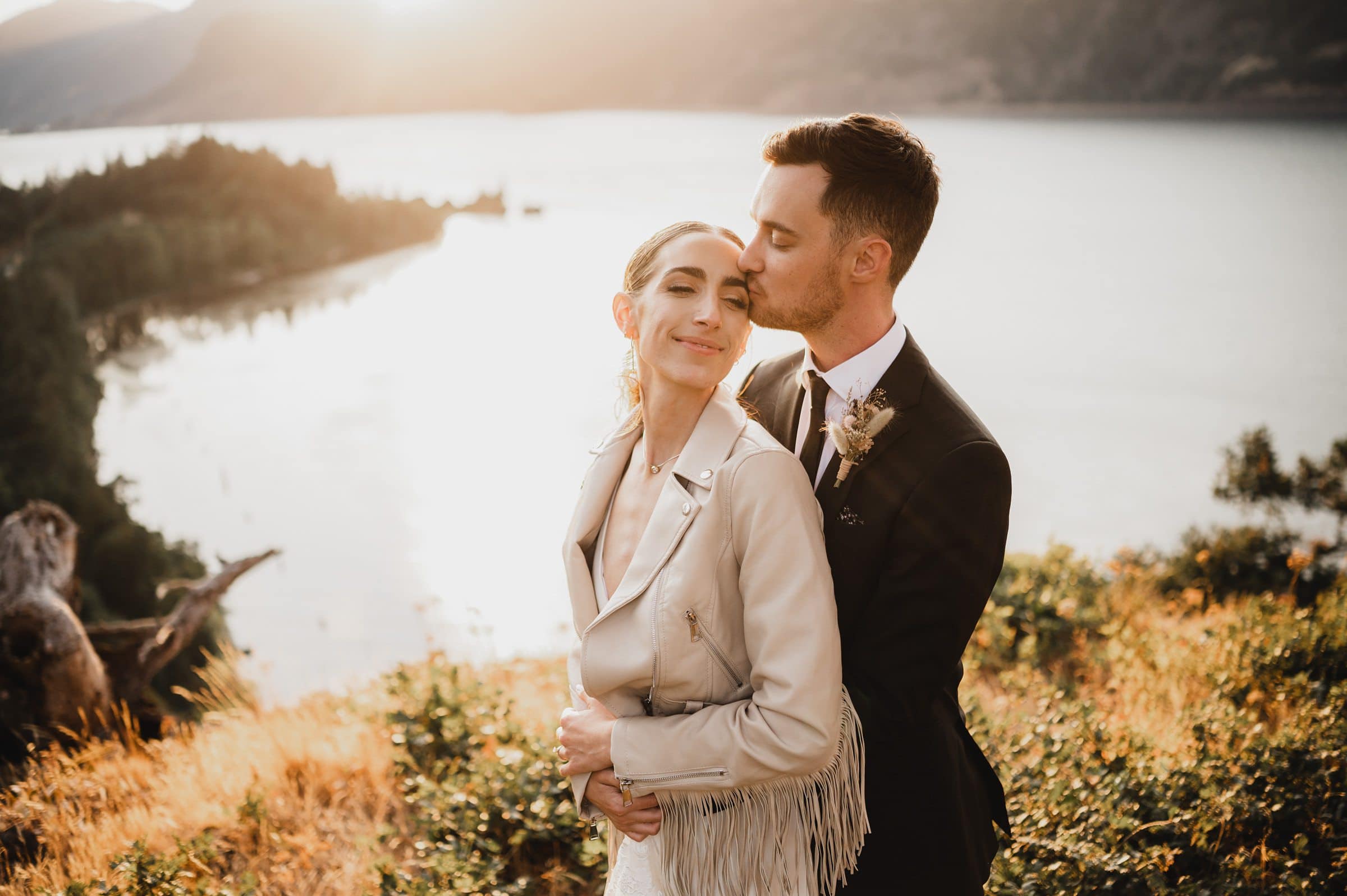 A couple embraces on cliffside in the Columbia River Gorge at sunset, with the groom kissing the brides head, capturing the moment in wedding photos at The Griffin House.