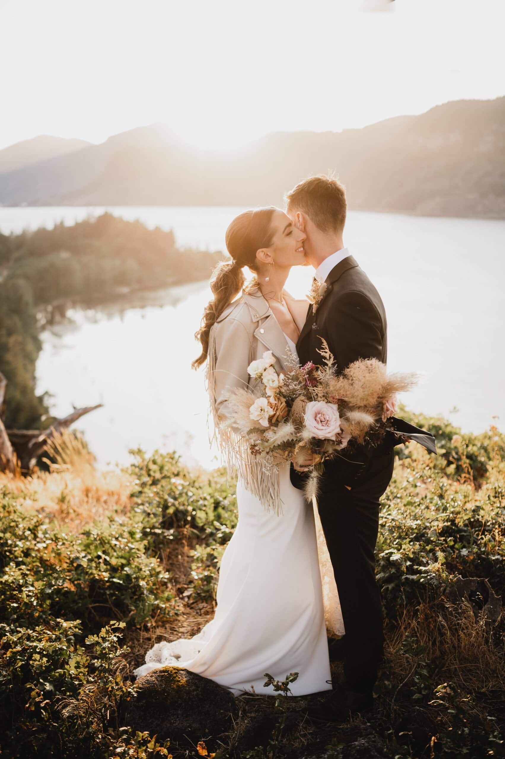 A couple in wedding attire embraces on a hillside with a scenic lake and mountain backdrop at sunset, capturing timeless wedding photos at The Griffin House.