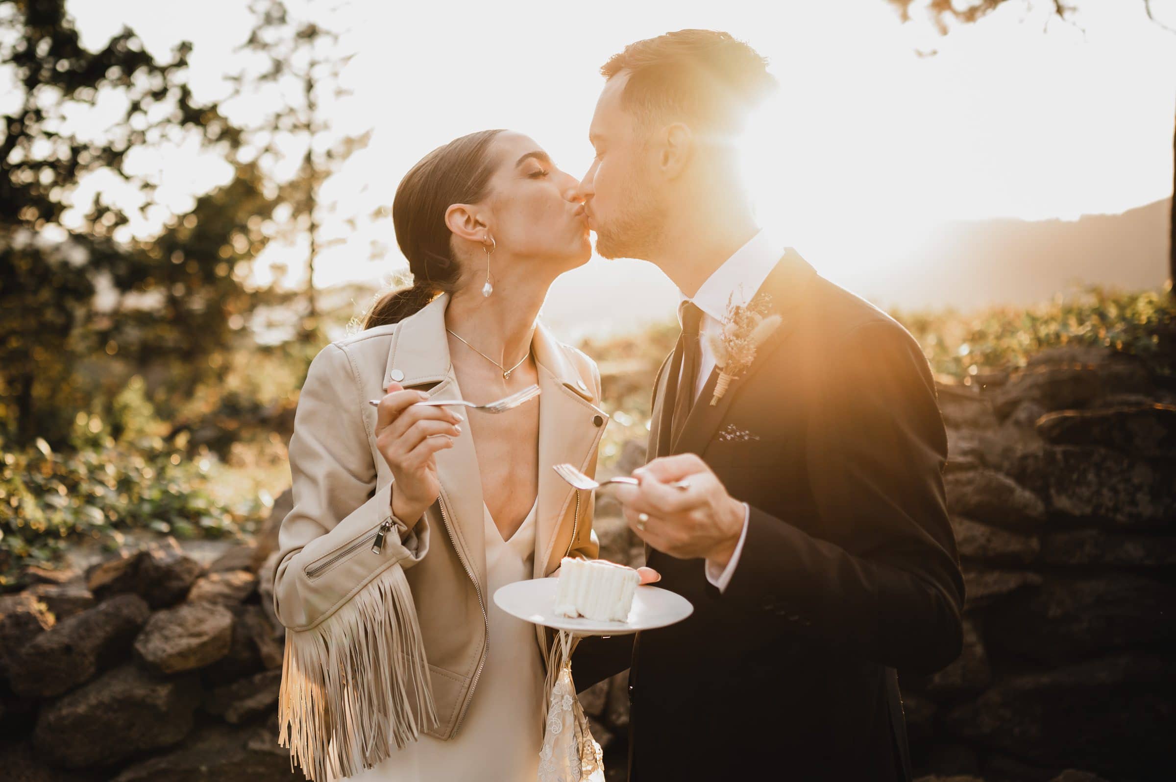 A couple shares a kiss at sunset, holding a slice of cake with forks in an outdoor setting, capturing the romance of their wedding photos at The Griffin House.