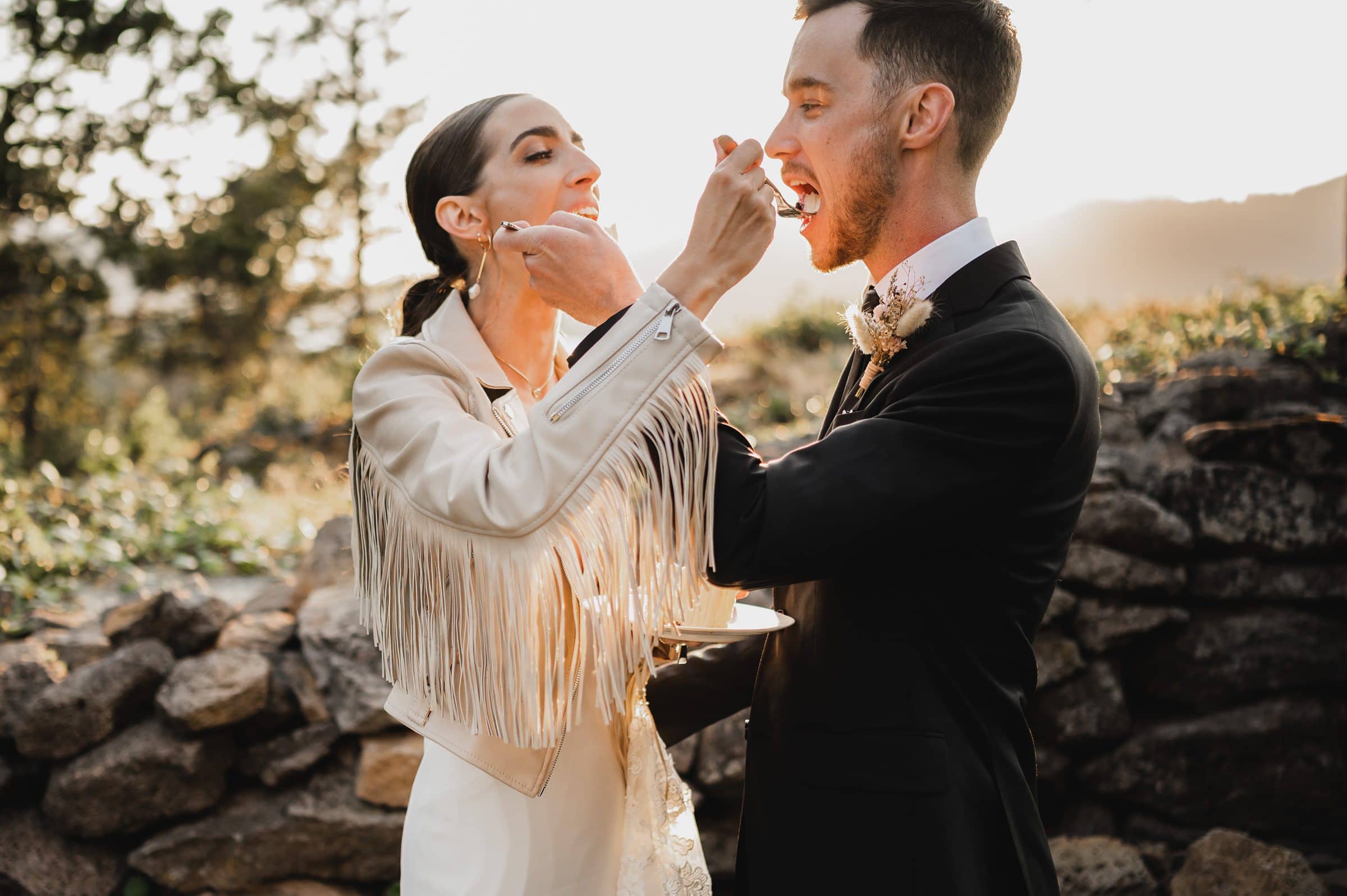 The bride and groom joyfully feed each other cake outdoors, surrounded by rocks and trees during a stunning sunset. Captured beautifully among nature, these wedding photos at The Griffin House create unforgettable memories.