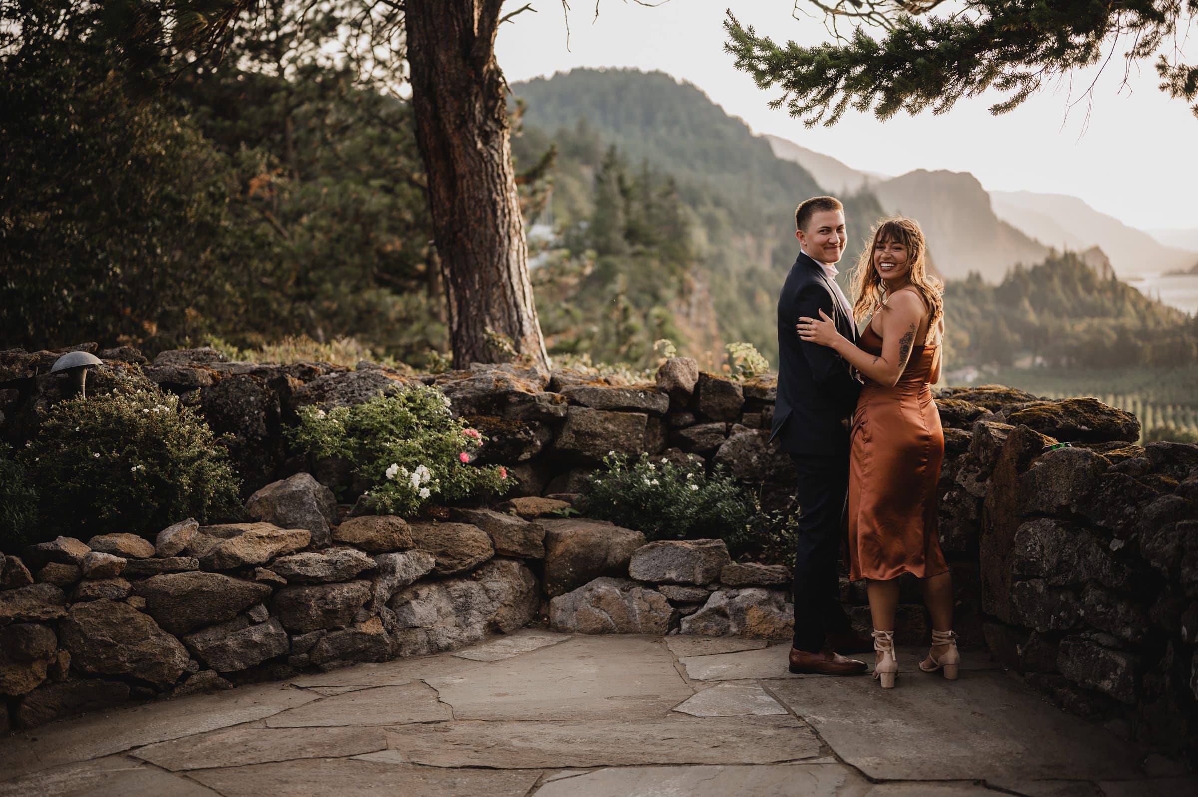 A couple smiling, standing by a stone wall with a scenic mountain view in the background, captures the joy of wedding photos at The Griffin House.