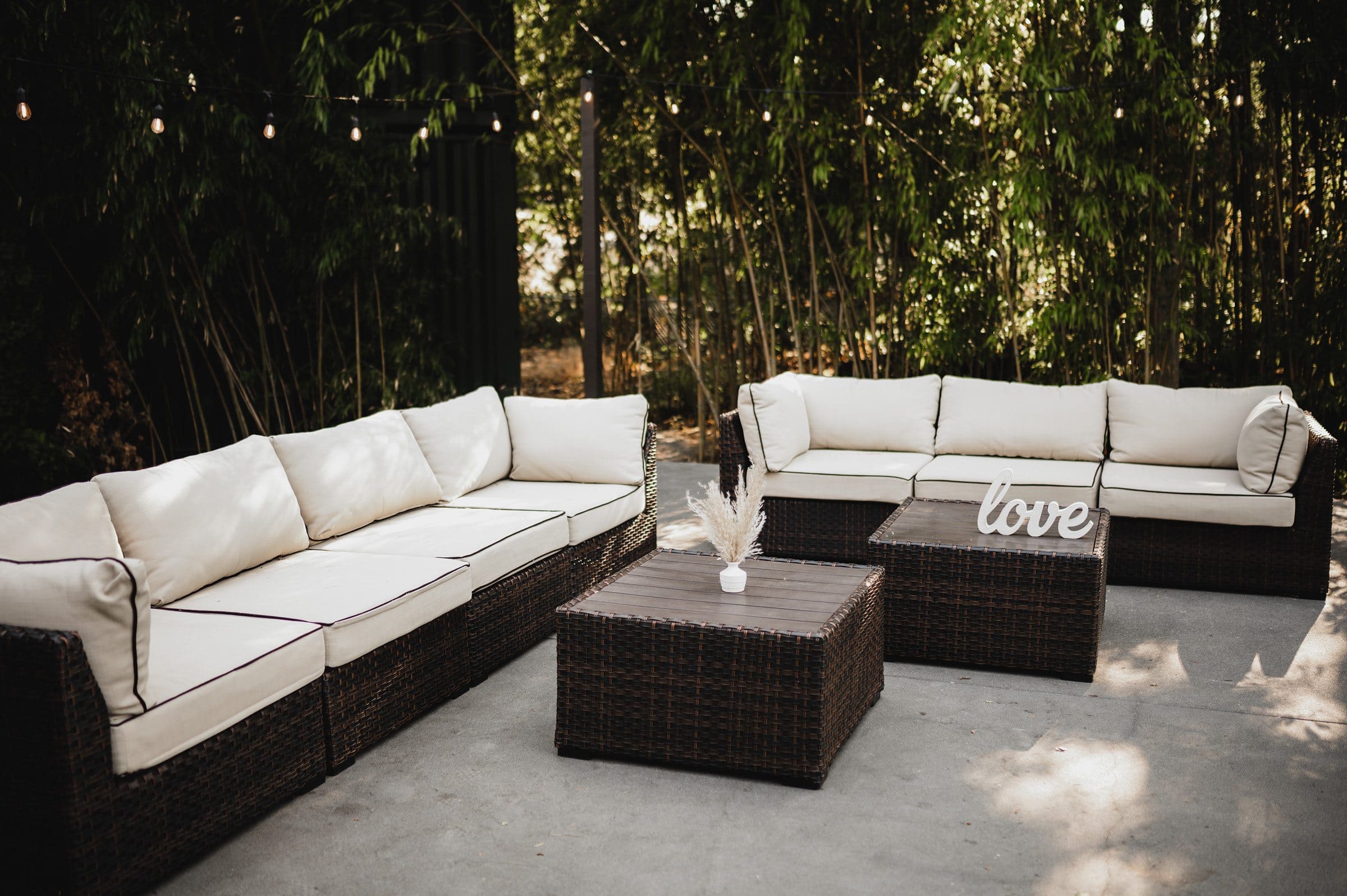 Outdoor seating area featuring white cushioned wicker sofas and a wooden table, surrounded by greenery and string lights—perfect for wedding photos at The Griffin House.