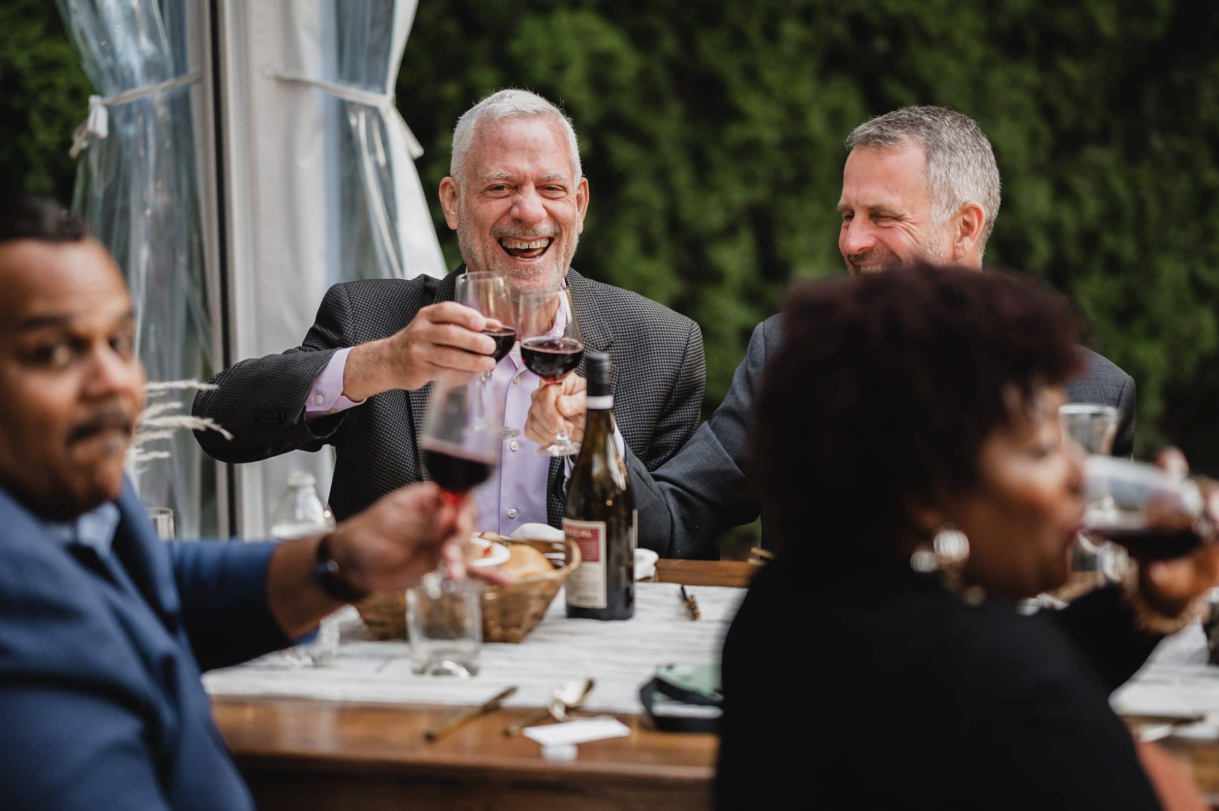 People joyfully toasting with wine glasses at an outdoor table, surrounded by lush greenery, capturing a moment akin to wedding photos at The Griffin House.