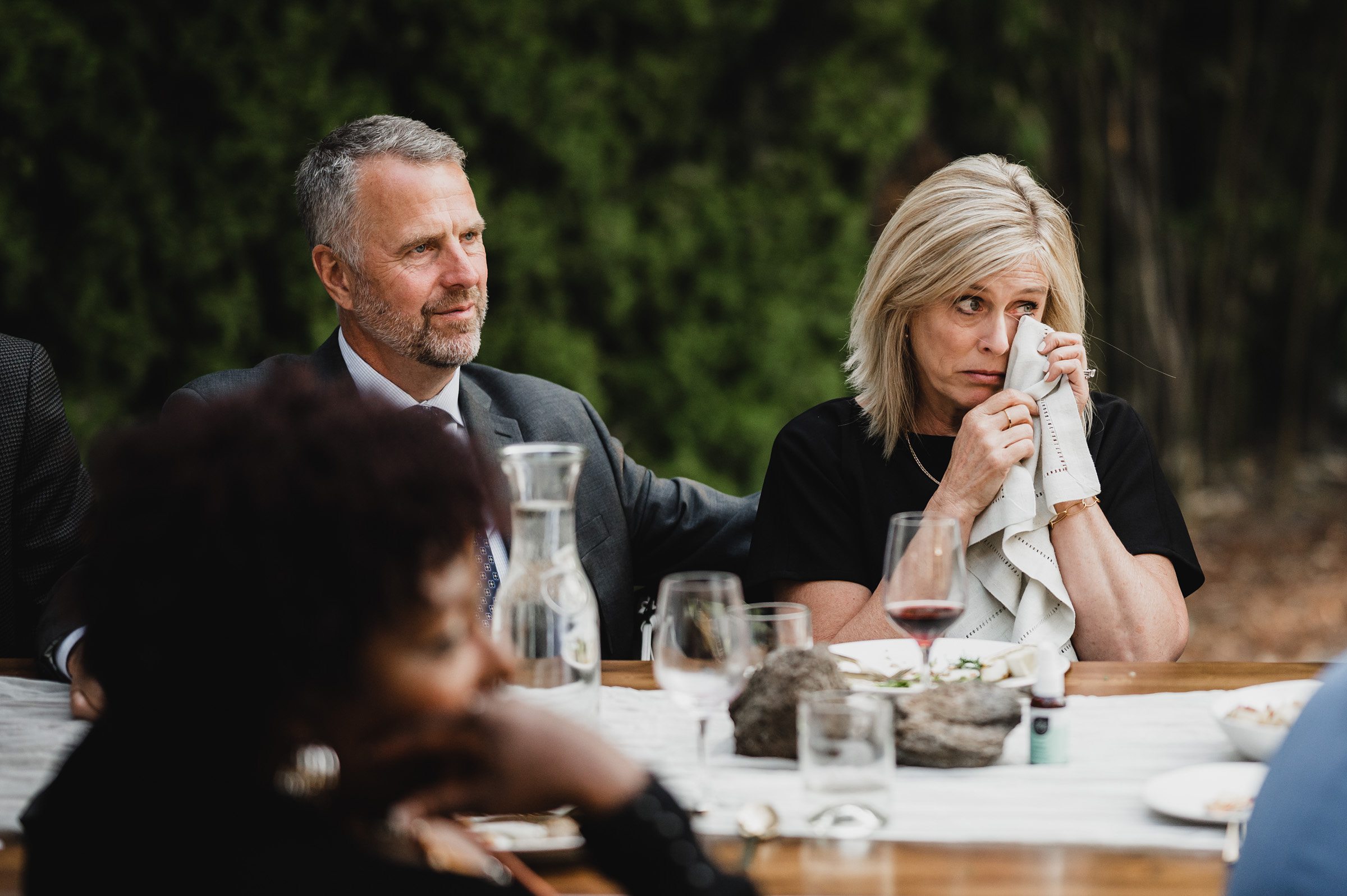 A man comforts a woman as she wipes her eyes with a napkin, seated at an outdoor table. Nearby, the backdrop of wedding photos at The Griffin House creates a picturesque setting that perfectly captures their tender moment.