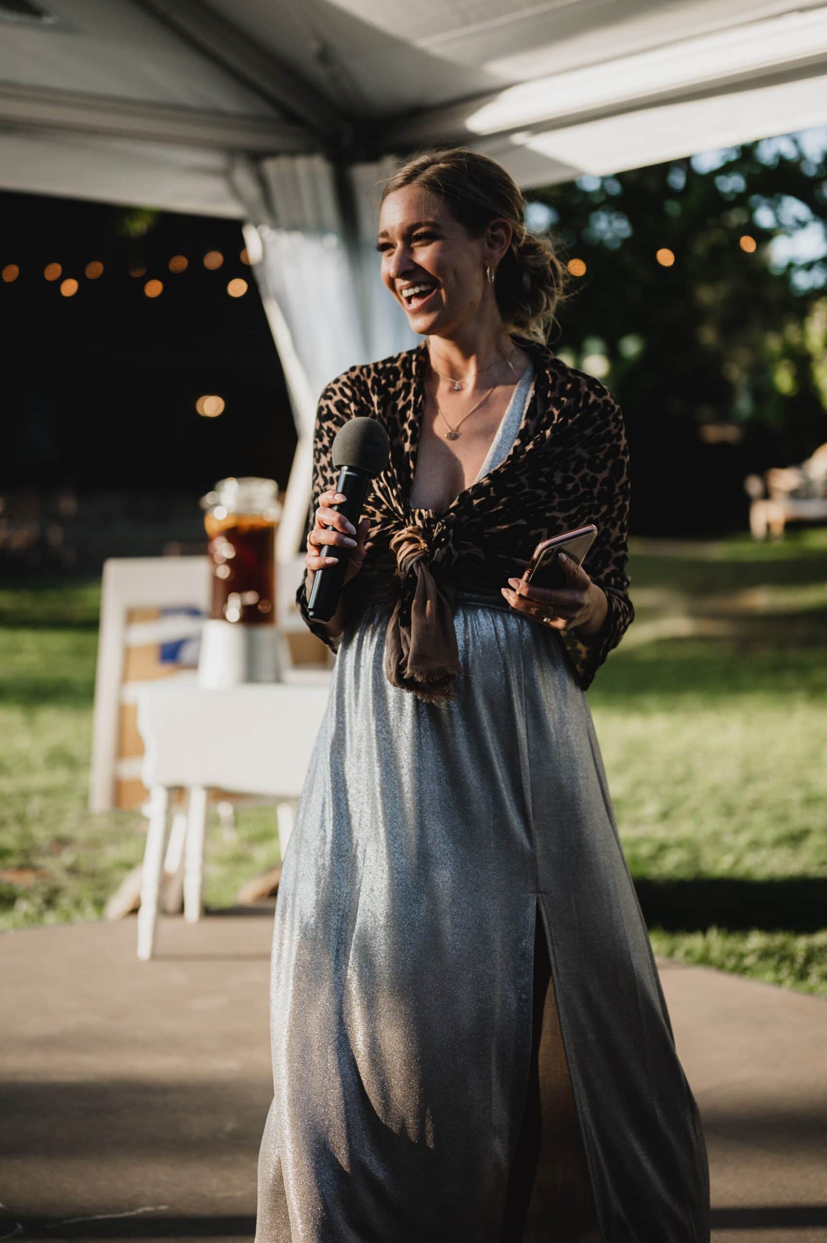 A woman in a silver dress and leopard-print top speaks into a microphone, smiling beneath the open sky. She adds a joyful touch to the enchanting atmosphere, perhaps capturing unforgettable moments akin to wedding photos at The Griffin House.