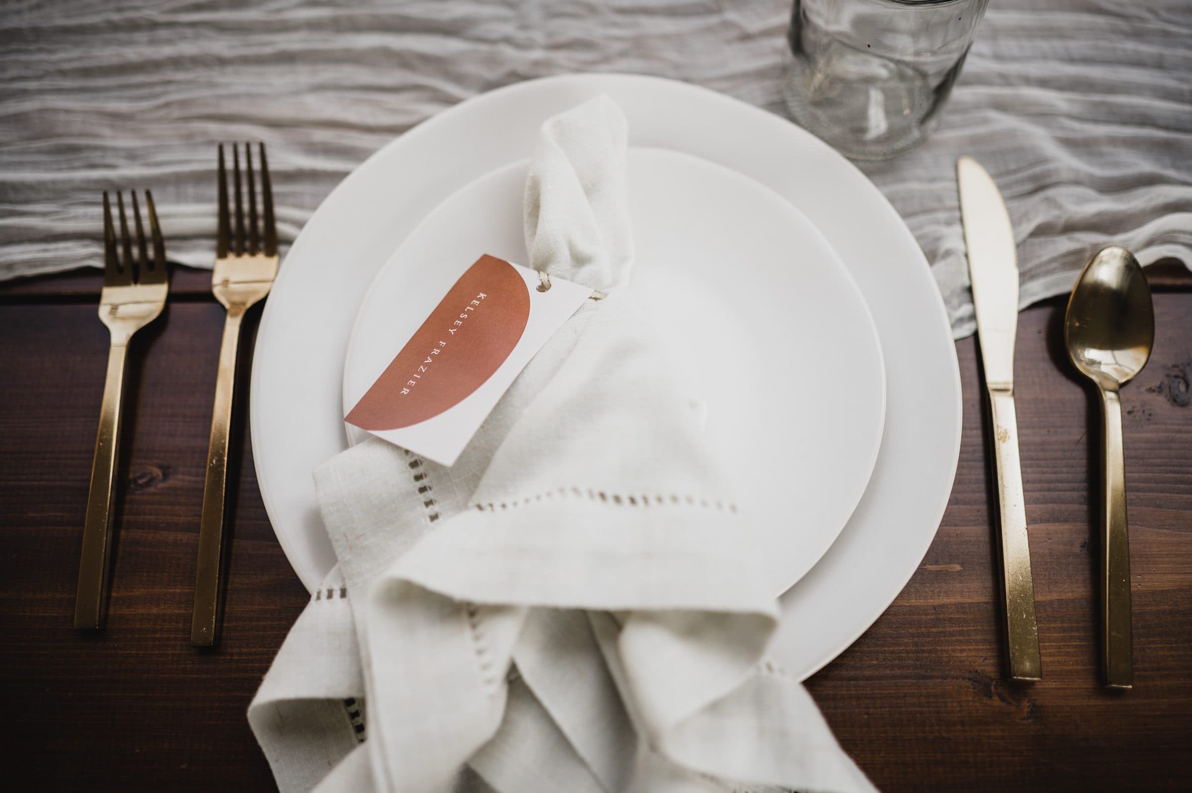 The elegant dining setup features gold cutlery, white plates, and a napkin on a wood table, complete with a glass and name card. Perfect for capturing stunning wedding photos at The Griffin House.