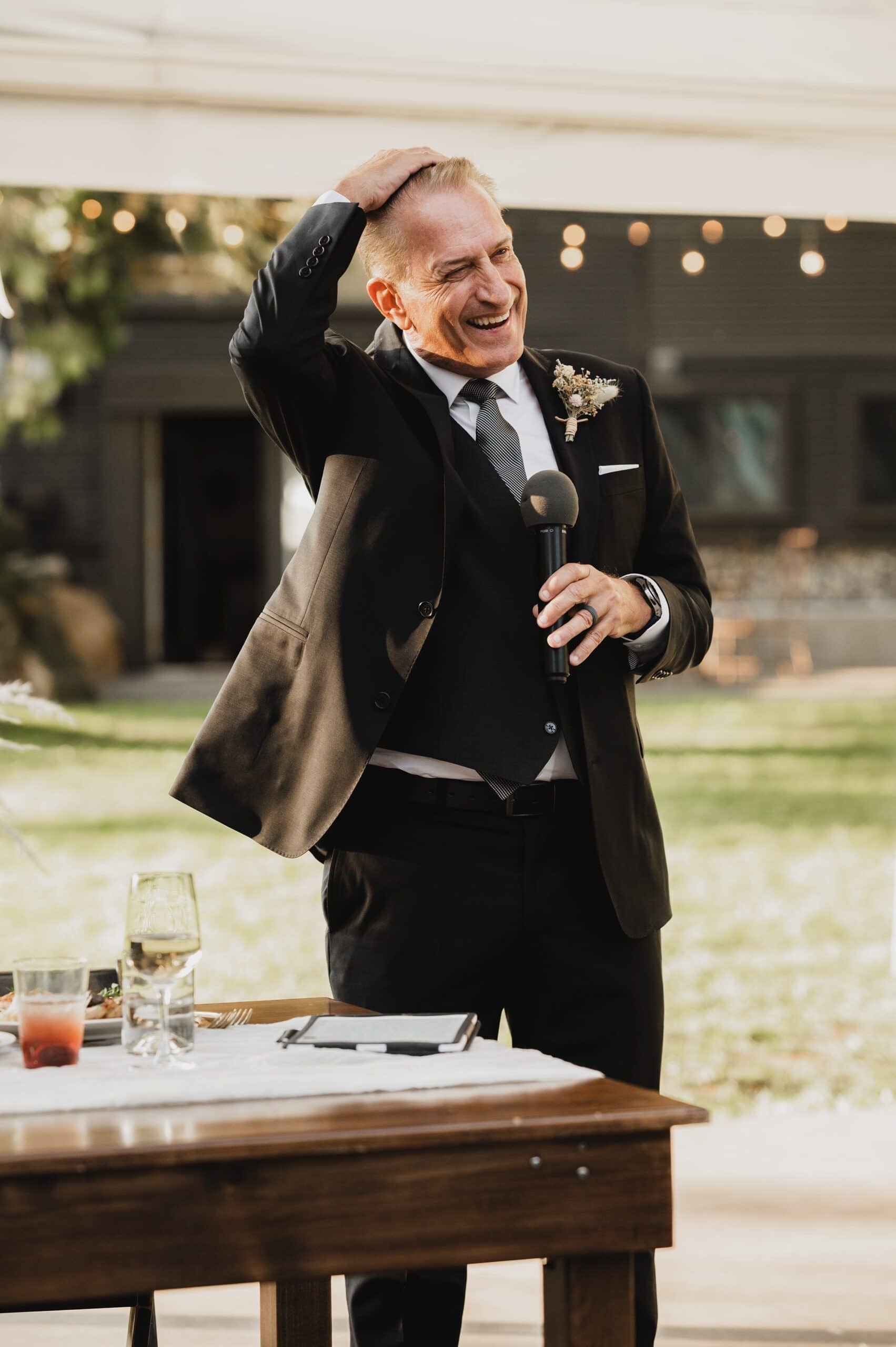 A man in a suit laughs while holding a microphone during an outdoor event at The Griffin House, standing next to a table with drinks.