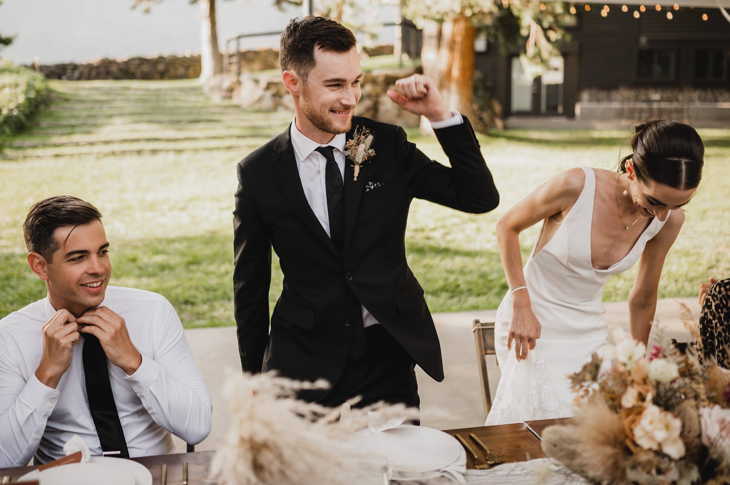 A smiling groom in a black suit gestures excitedly at an outdoor reception at The Griffin House, with the bride in a white dress nearby.