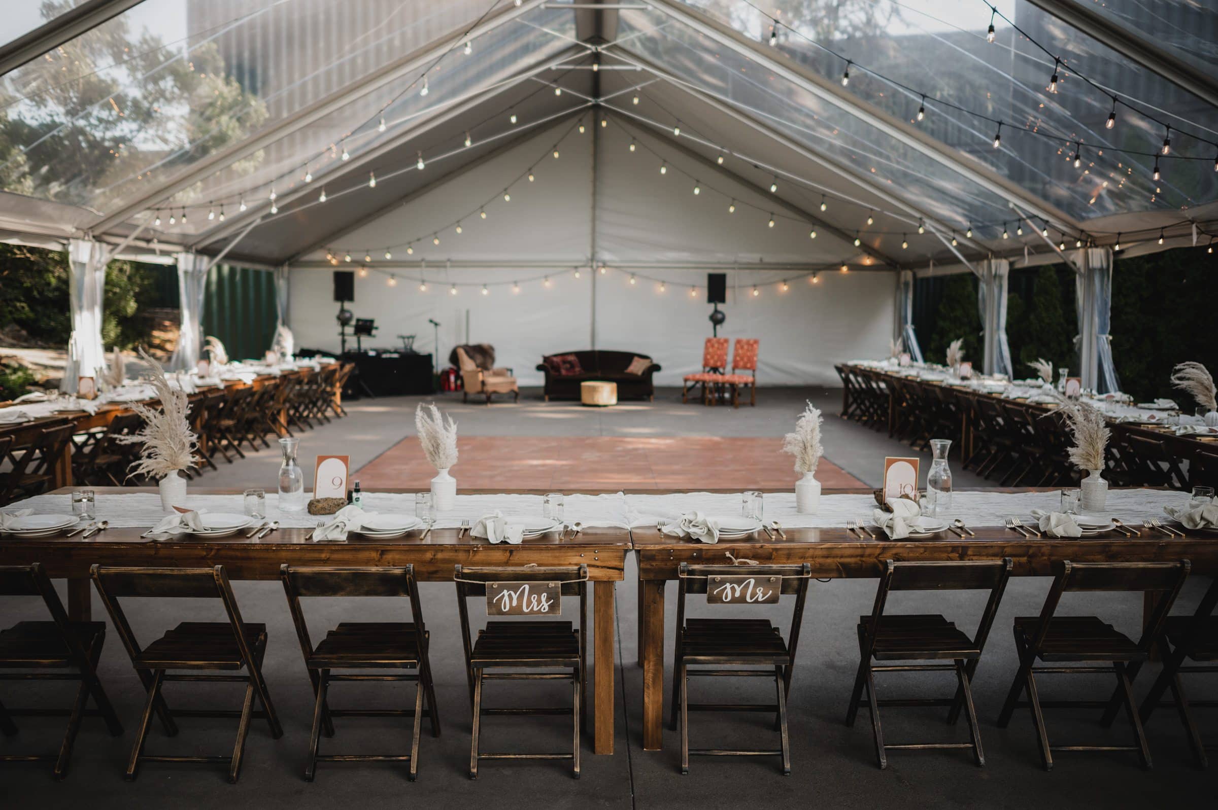 Experience a magical wedding reception setup under a clear tent adorned with string lights, featuring long wooden tables and chairs beside the dance floor. Capture unforgettable wedding photos at The Griffin House for a truly enchanting celebration.