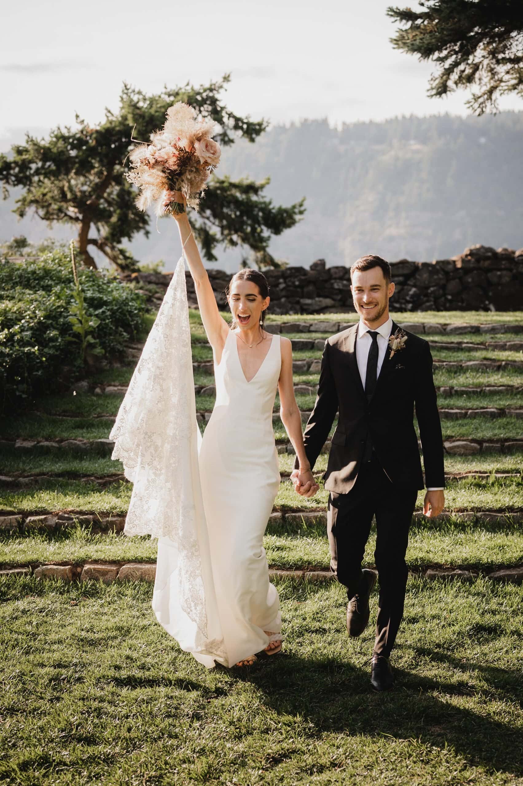 The bride and groom walk hand in hand outdoors, with the bride holding her bouquet and veil aloft. In the background of their wedding photos at The Griffin House, lush grass and towering trees create a picturesque scene.