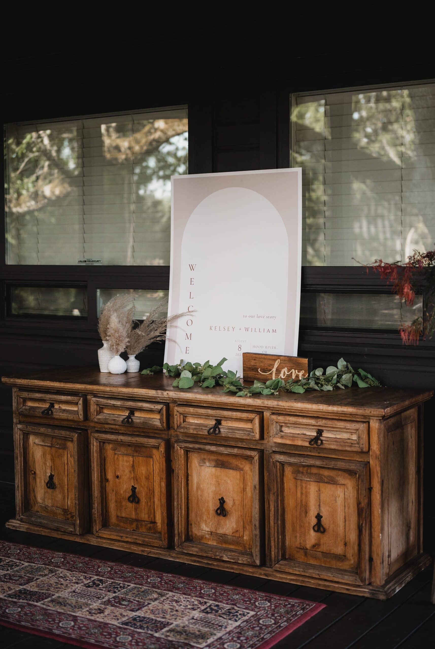A wooden sideboard adorned with greenery, pampas grass, and a Welcome wedding sign creates a charming scene in a sunlit room with large windows, ideal for capturing stunning wedding photos at The Griffin House.