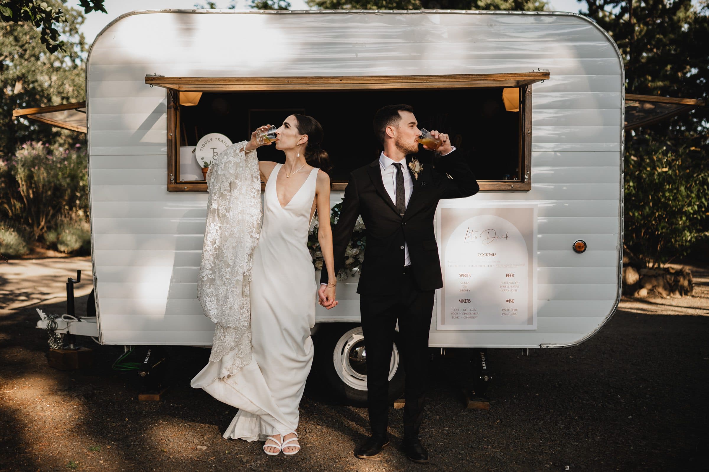 The bride and groom sip drinks, leaning against a vintage trailer outside, holding hands with greenery in the background, capturing a perfect moment for their wedding photos at The Griffin House.