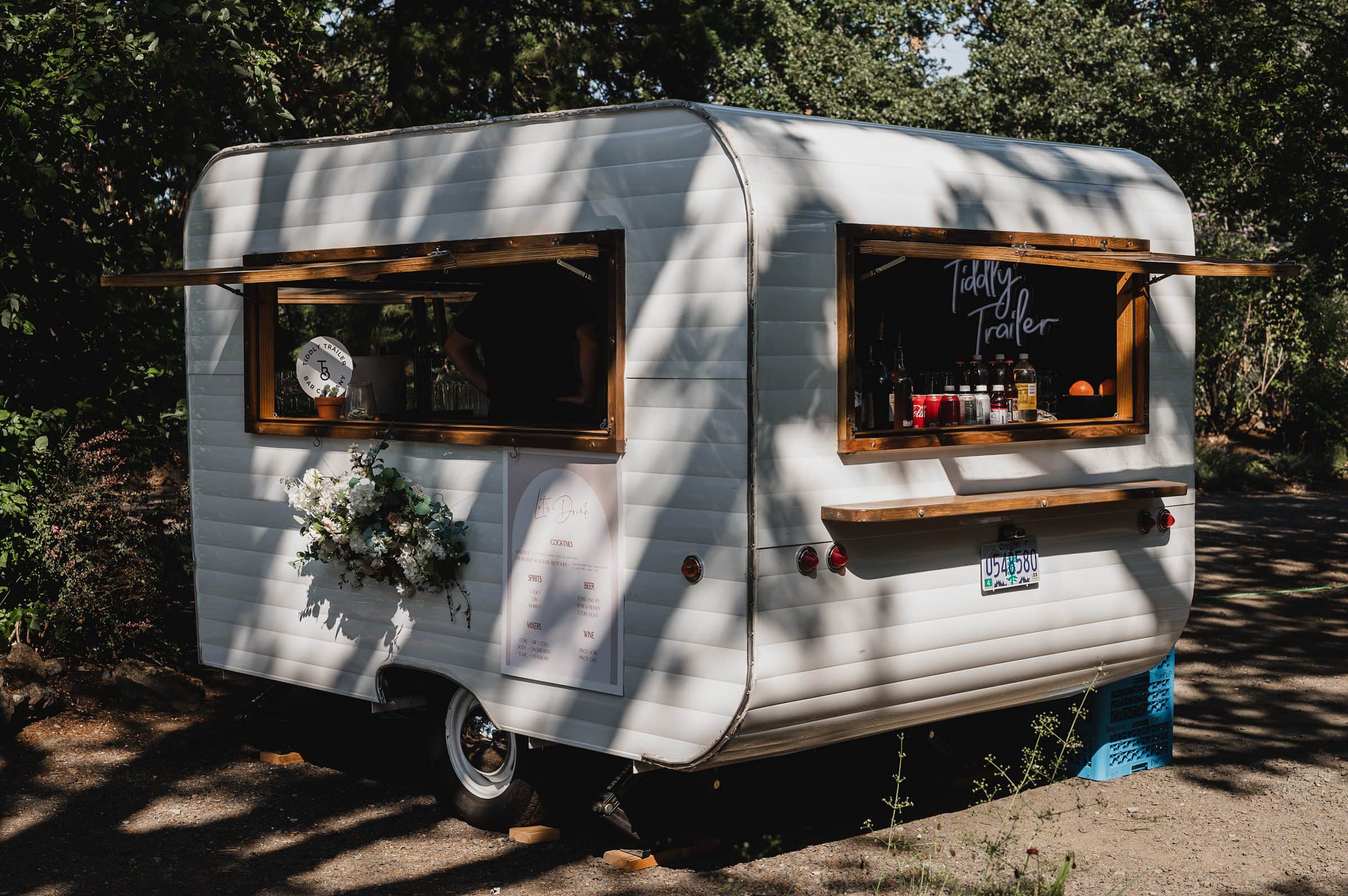A small white trailer with open windows is parked among trees, adorned with signs and flowers—perfect for capturing wedding photos at The Griffin House.