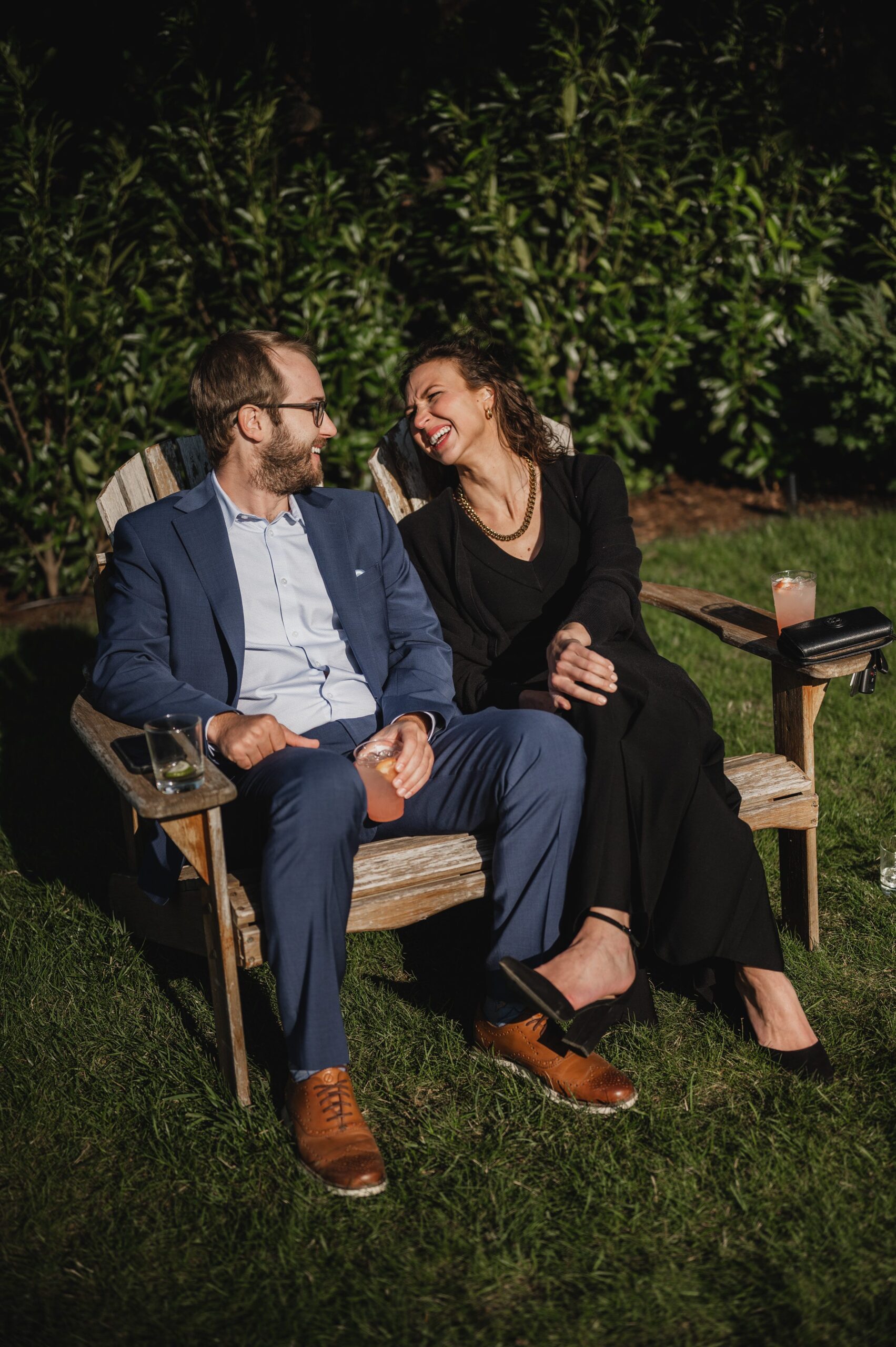 A man and woman sit on a wooden bench outdoors at The Griffin House, laughing and holding drinks, as they pose for wedding photos.