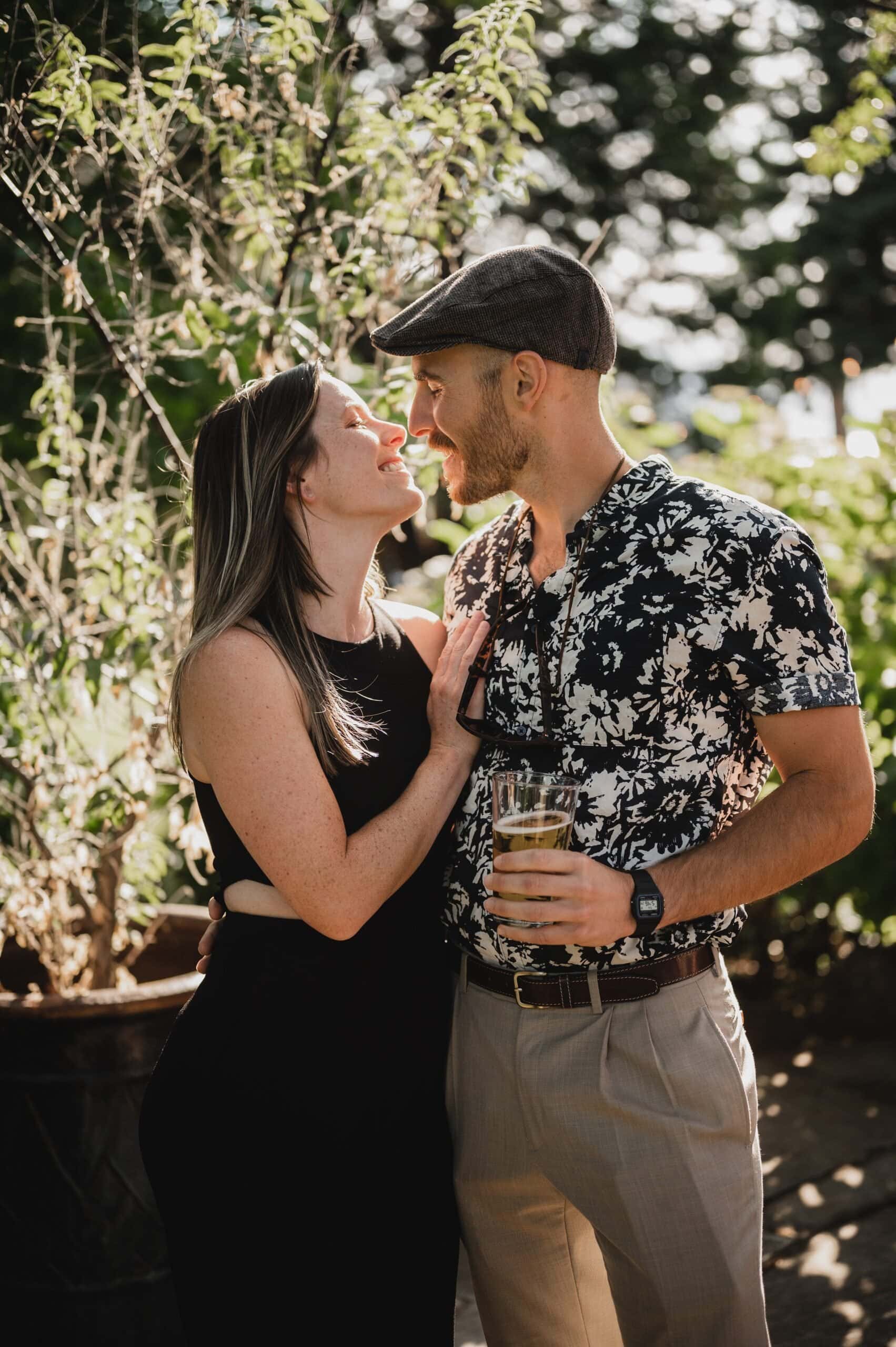 A smiling couple outdoors, the woman in a black dress and the man in a patterned shirt and cap, holding a drink during wedding photos at The Griffin House.
