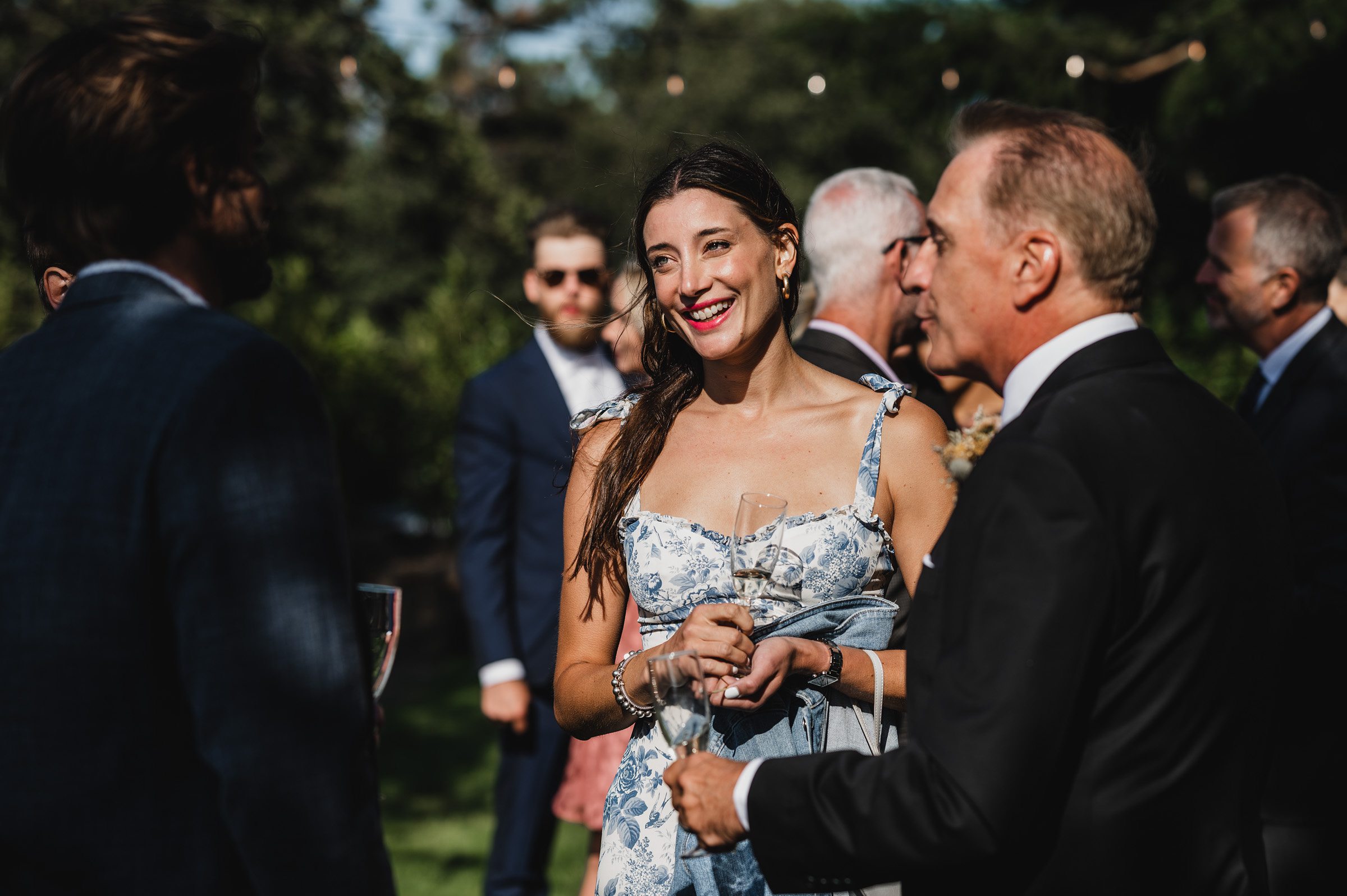 A woman in a floral dress holding a drink smiles at an outdoor gathering, surrounded by people in formal attire. The event captures the joy of wedding photos at The Griffin House, where everyone is dressed to impress.