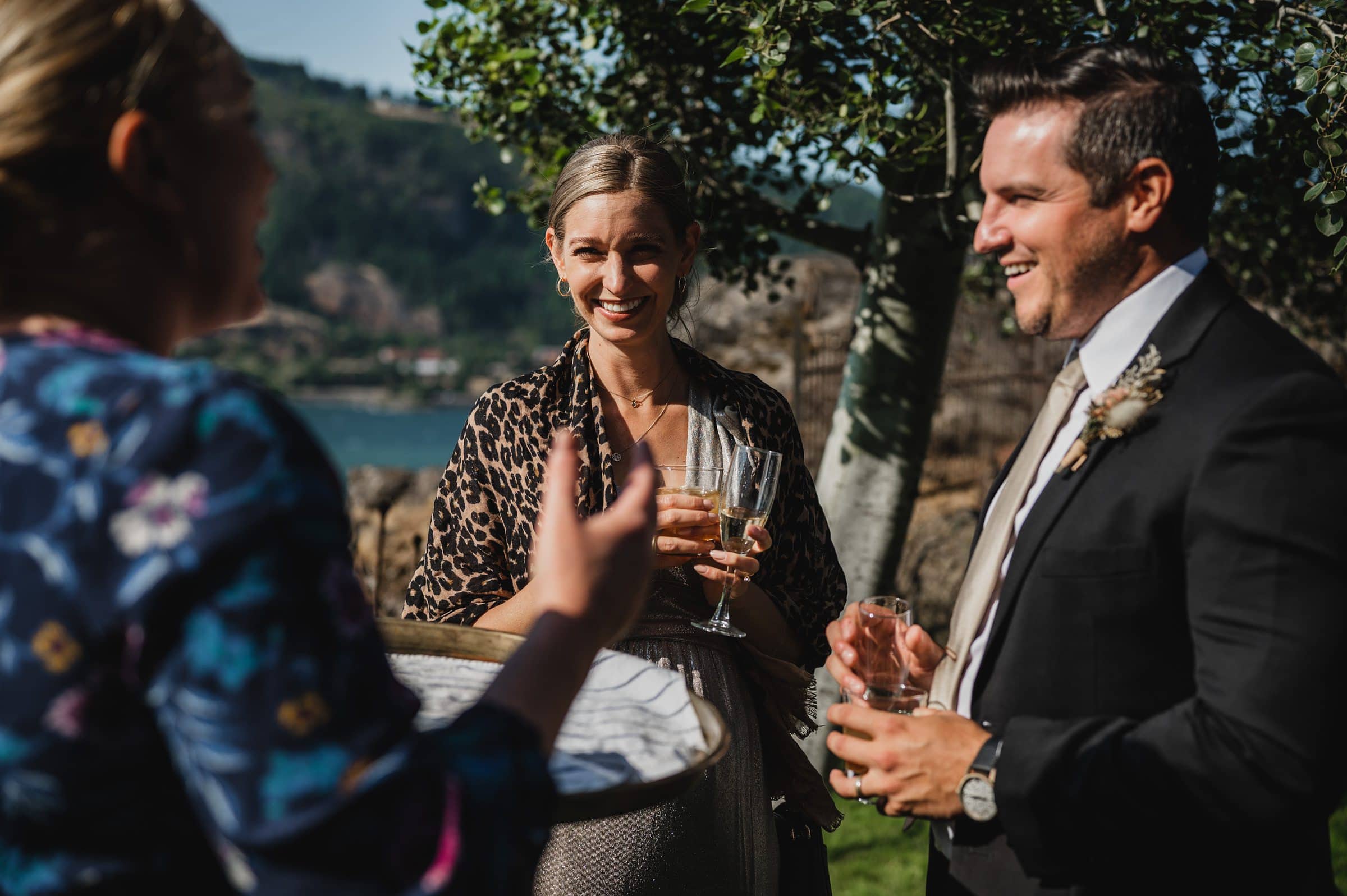 Three people smiling and chatting outdoors, holding drinks, with greenery and a body of water in the background—perfect for wedding photos at The Griffin House.
