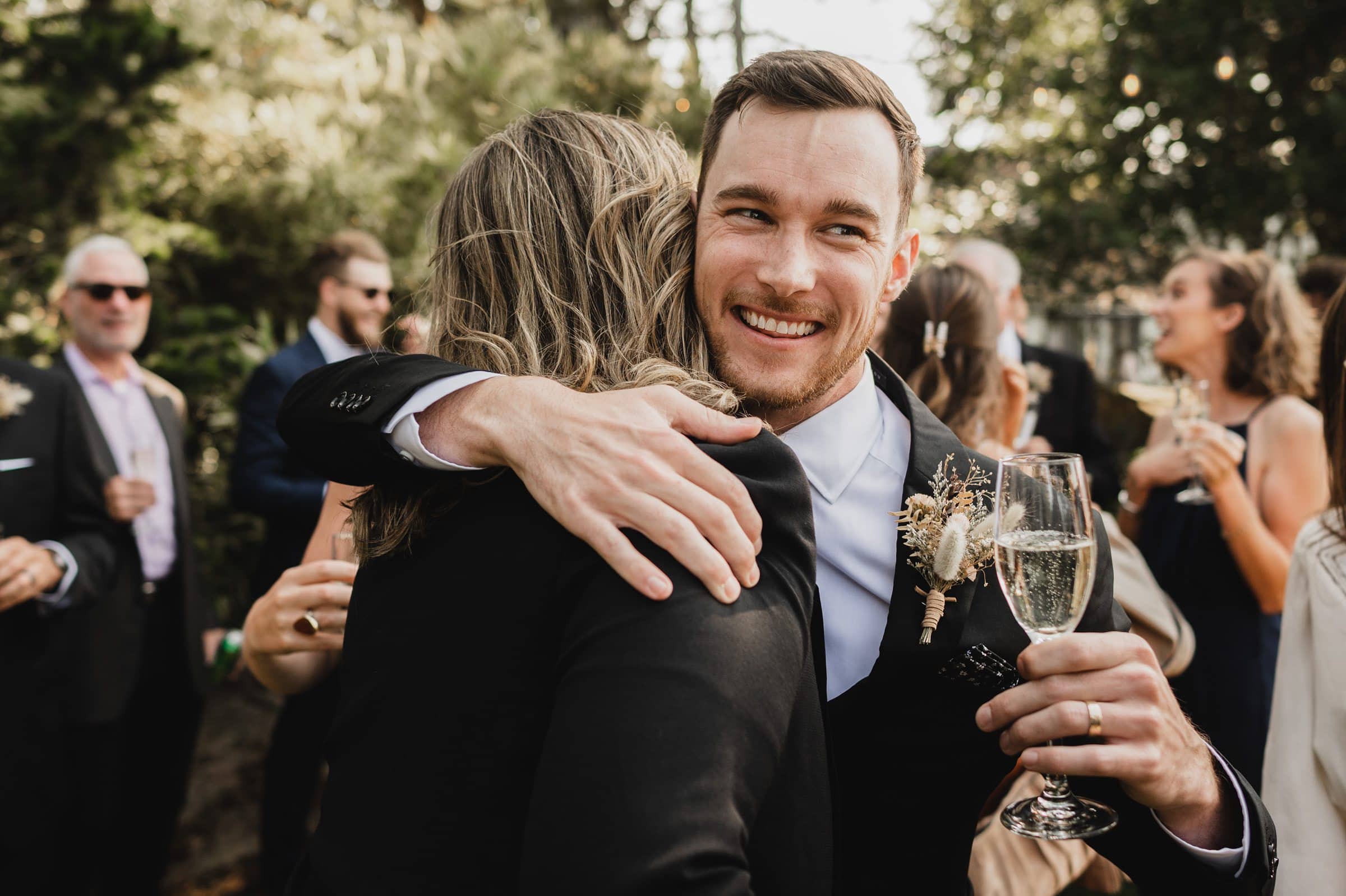 Man in a suit hugging someone at an outdoor event, smiling and holding a glass of champagne, capturing joyful wedding photos at The Griffin House.