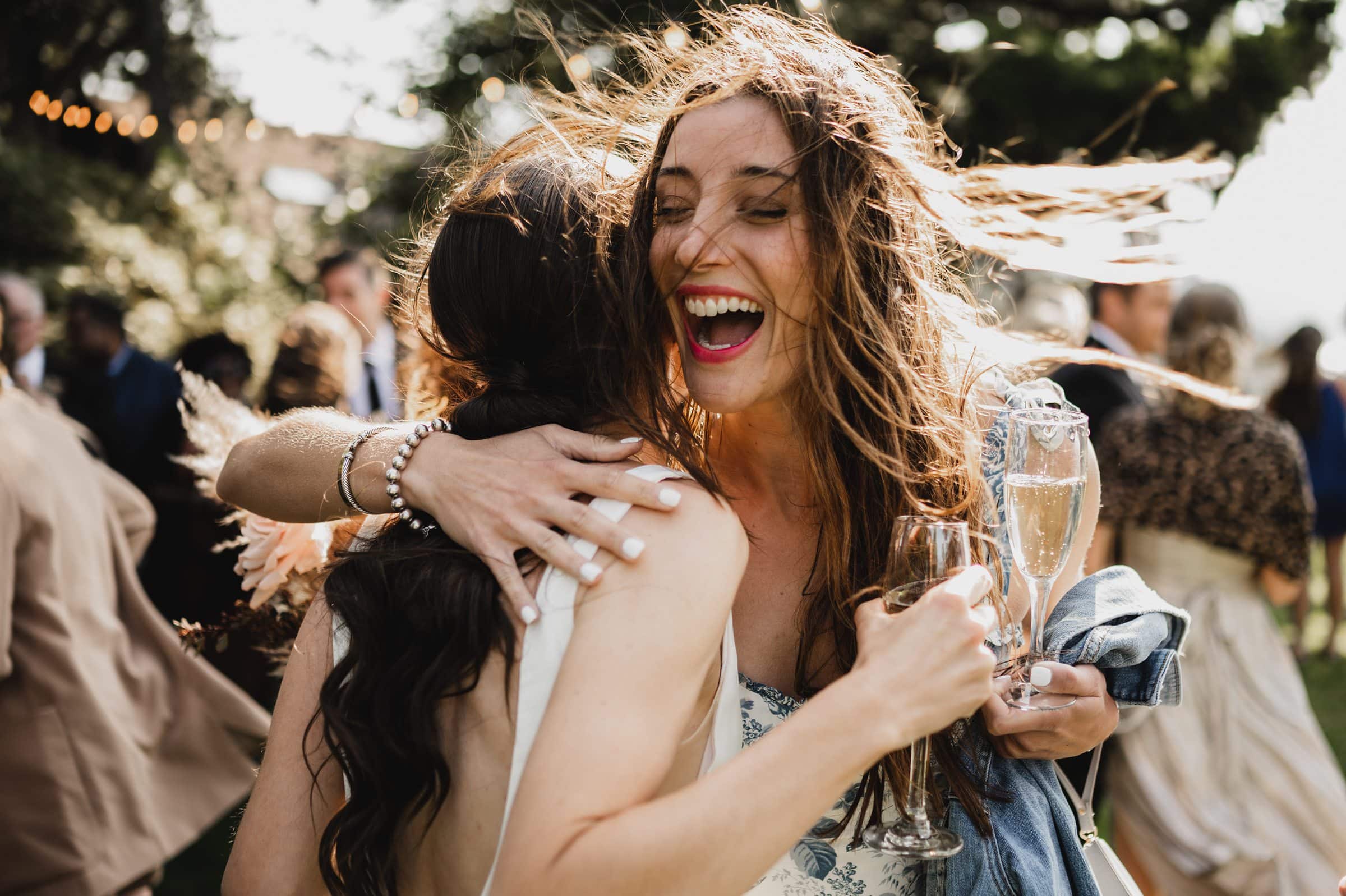 Two women celebrating outdoors, hugging and laughing, with one holding a champagne glass. The wind playfully lifts their hair in this joyful moment, reminiscent of wedding photos at The Griffin House.