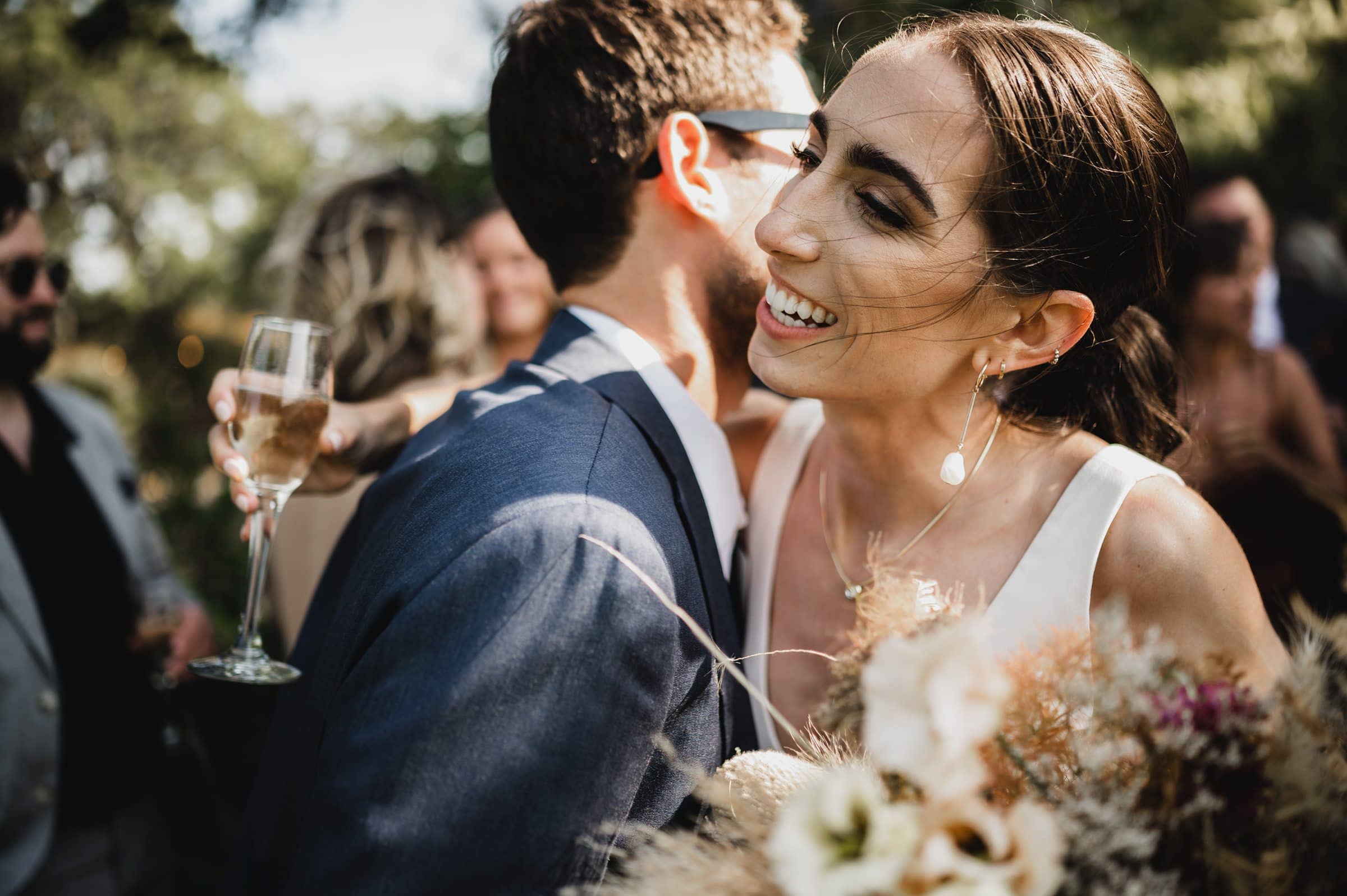 A woman in a white dress smiles, embracing a man in a suit as she holds a bouquet and champagne flute during wedding photos at The Griffin House.
