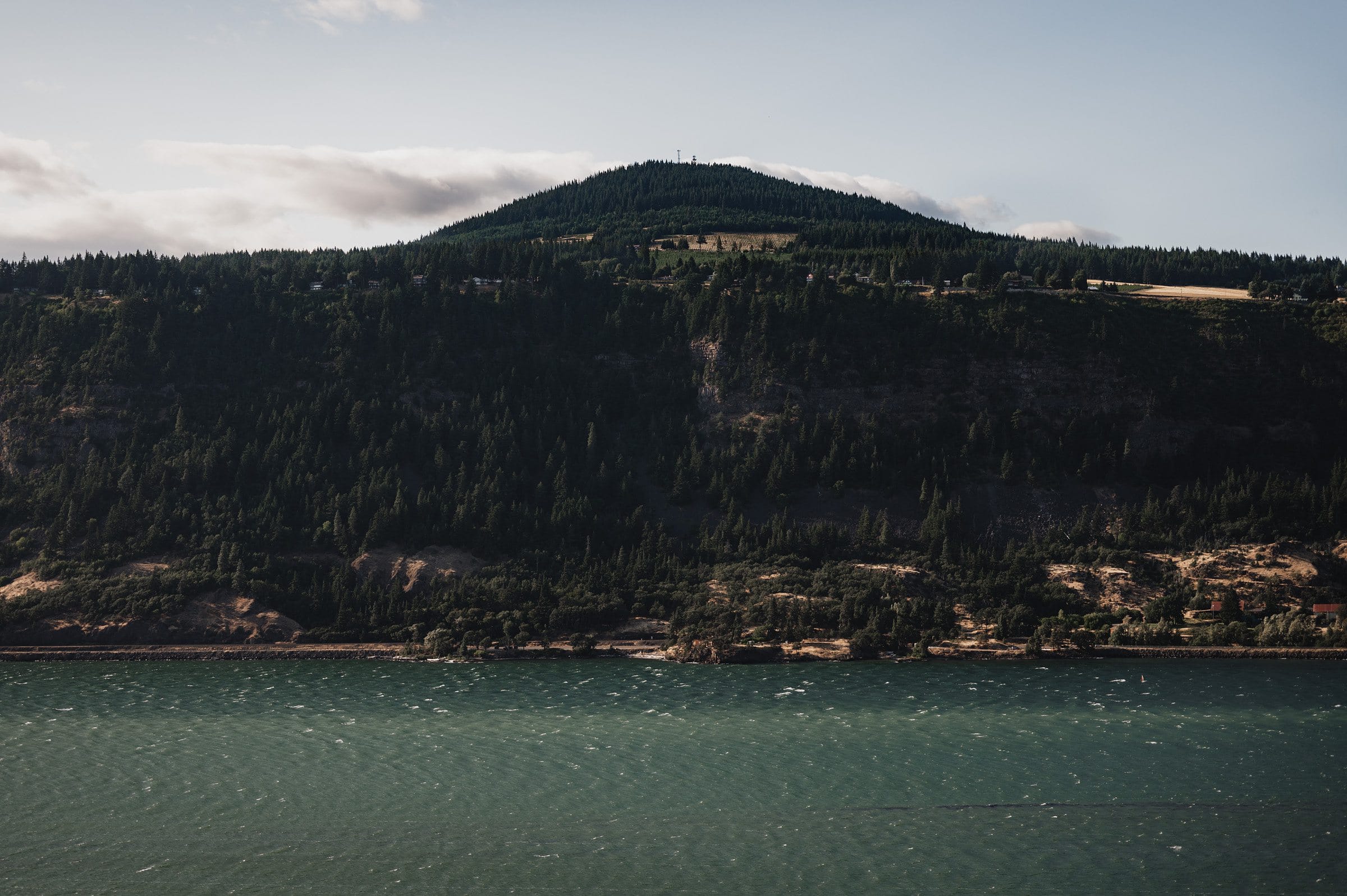 A forested hill with a few structures on top, surrounded by a river under a partly cloudy sky, creates the perfect backdrop for capturing wedding photos at The Griffin House.