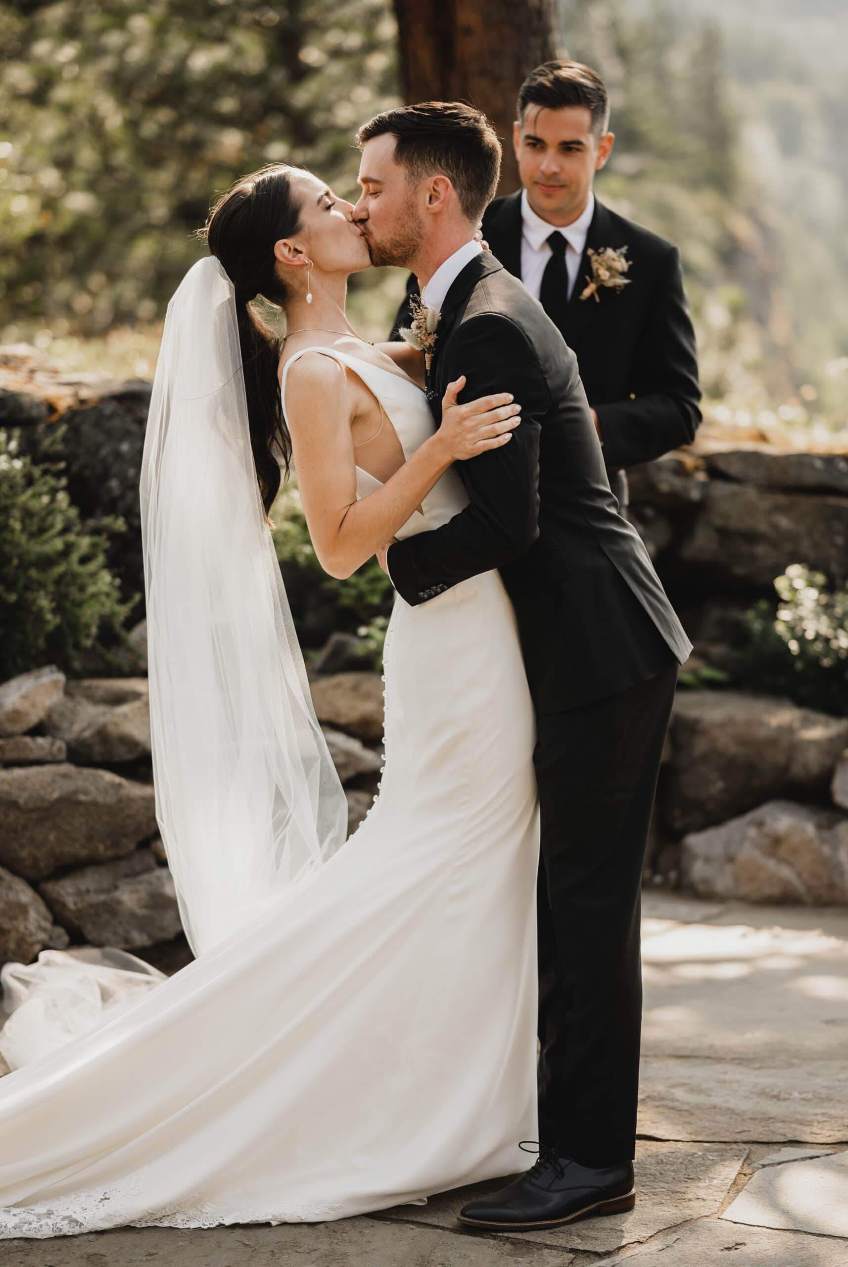 The bride and groom share a kiss at their outdoor wedding ceremony, with the officiant standing behind them. This moment is beautifully captured among the stunning surroundings of The Griffin House.