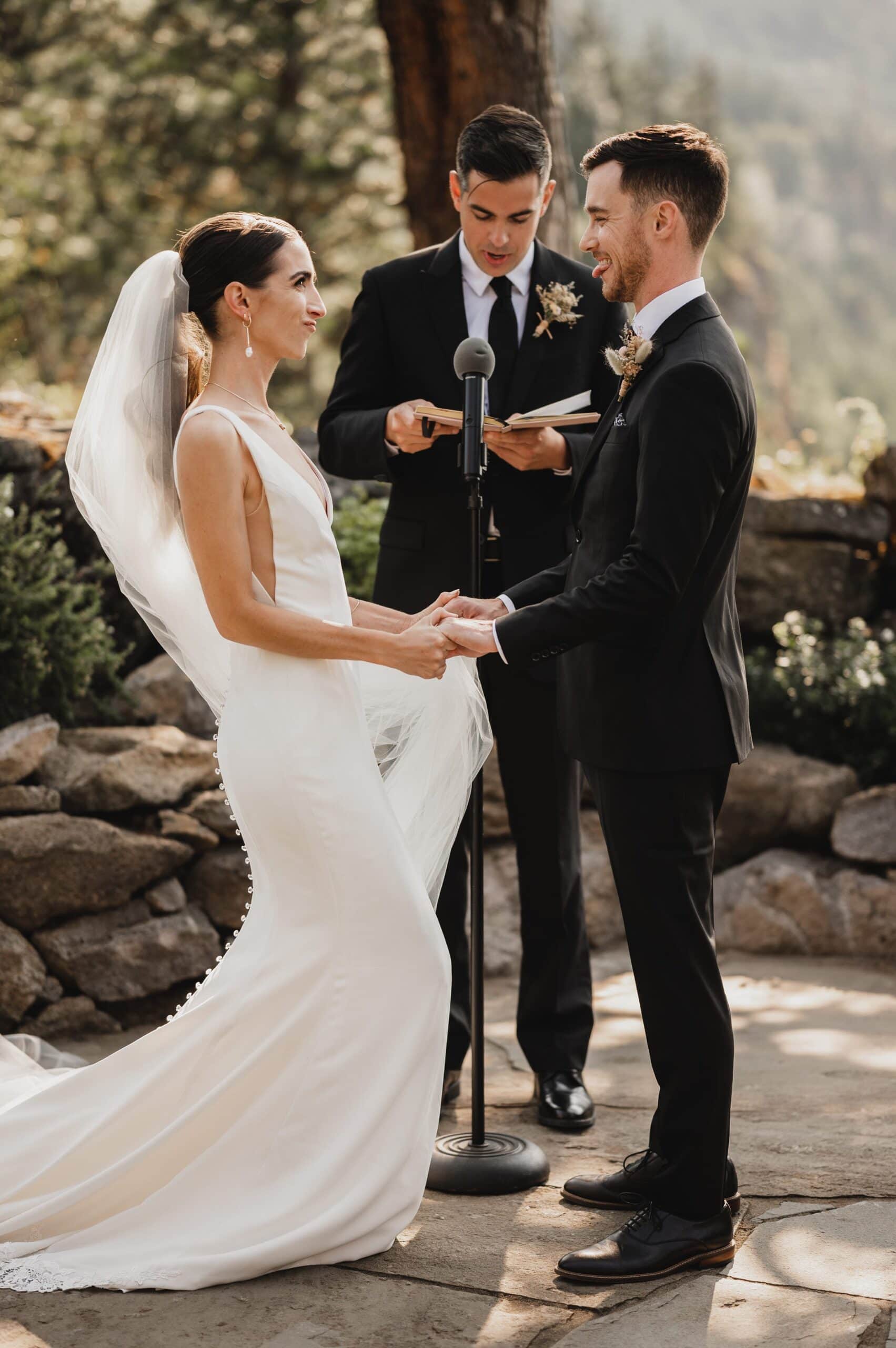 The bride and groom hold hands during their outdoor wedding ceremony at The Griffin House, as the officiant reads from a book.