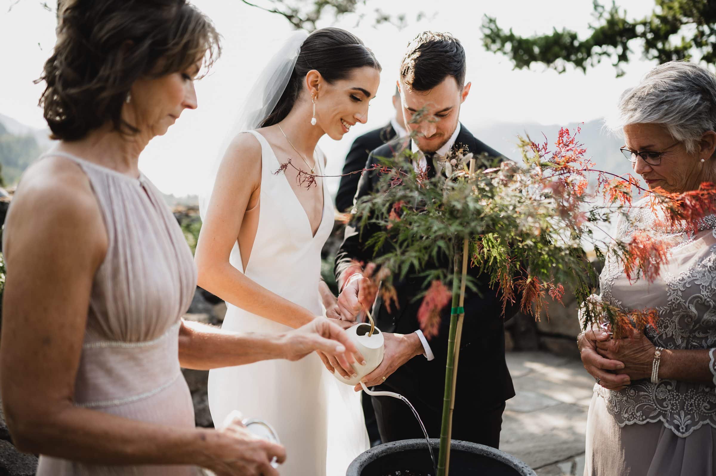During a sunny ceremony, a bride and groom, along with two women, plant a tree outdoors. Its the perfect moment for wedding photos at The Griffin House.