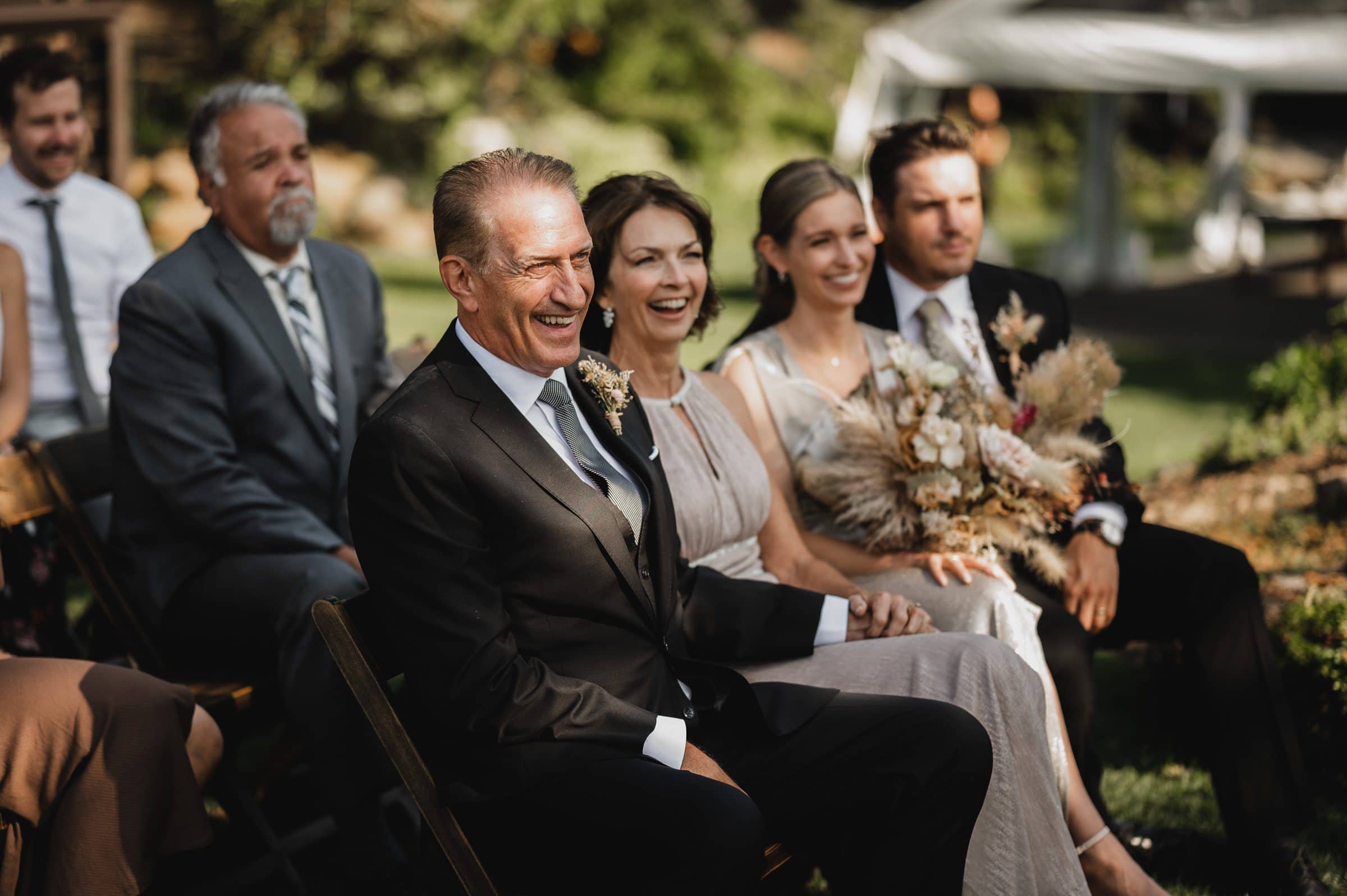 Guests at the wedding, all smiles as they sit outdoors with lush foliage and a tent in the background, make for stunning wedding photos at The Griffin House.