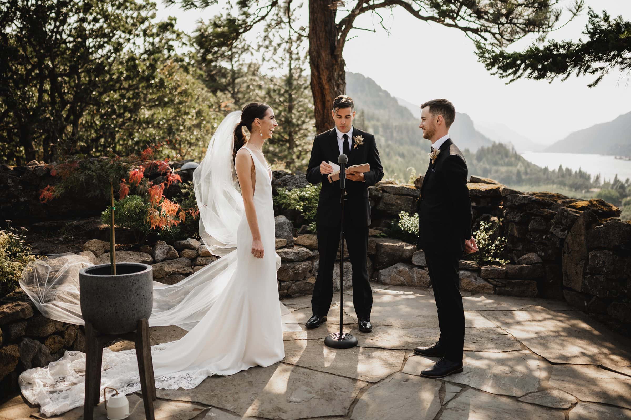 A couple stands facing each other during an outdoor wedding ceremony at The Griffin House, with a scenic view of mountains and a lake in the background.