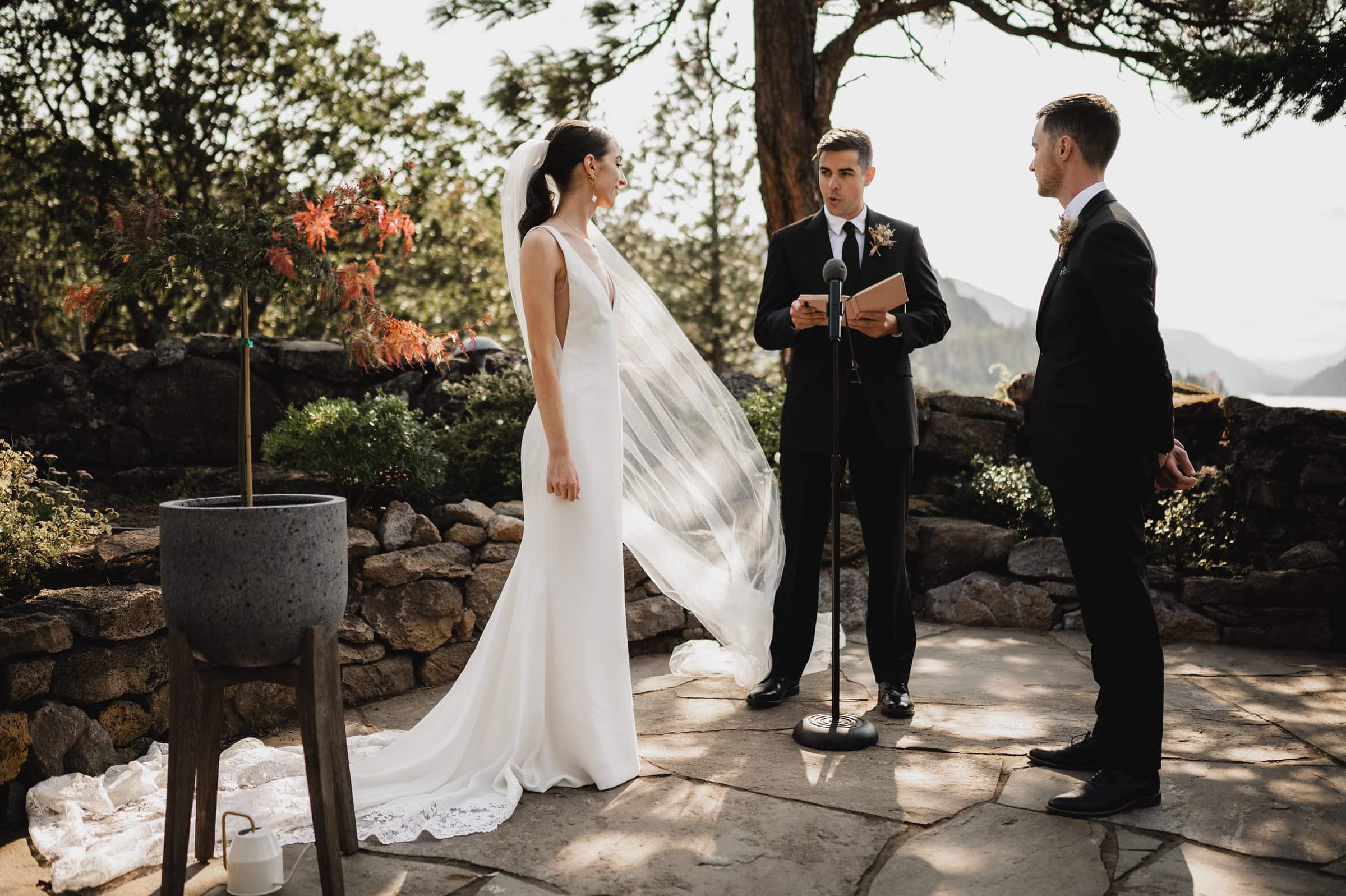 A bride and groom stand with an officiant outdoors during their wedding ceremony, surrounded by greenery at The Griffin House.